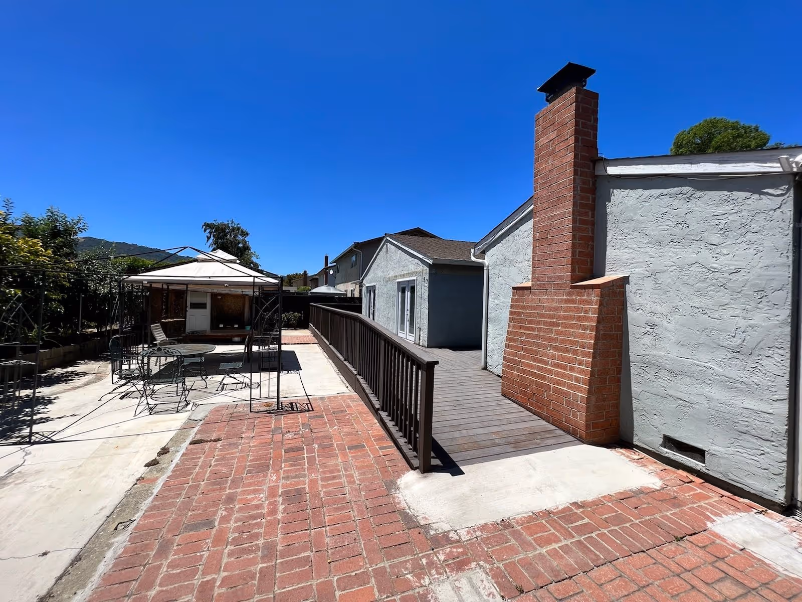 Sunny outdoor courtyard with brick patio, seating under a canopy beside a light gray building.