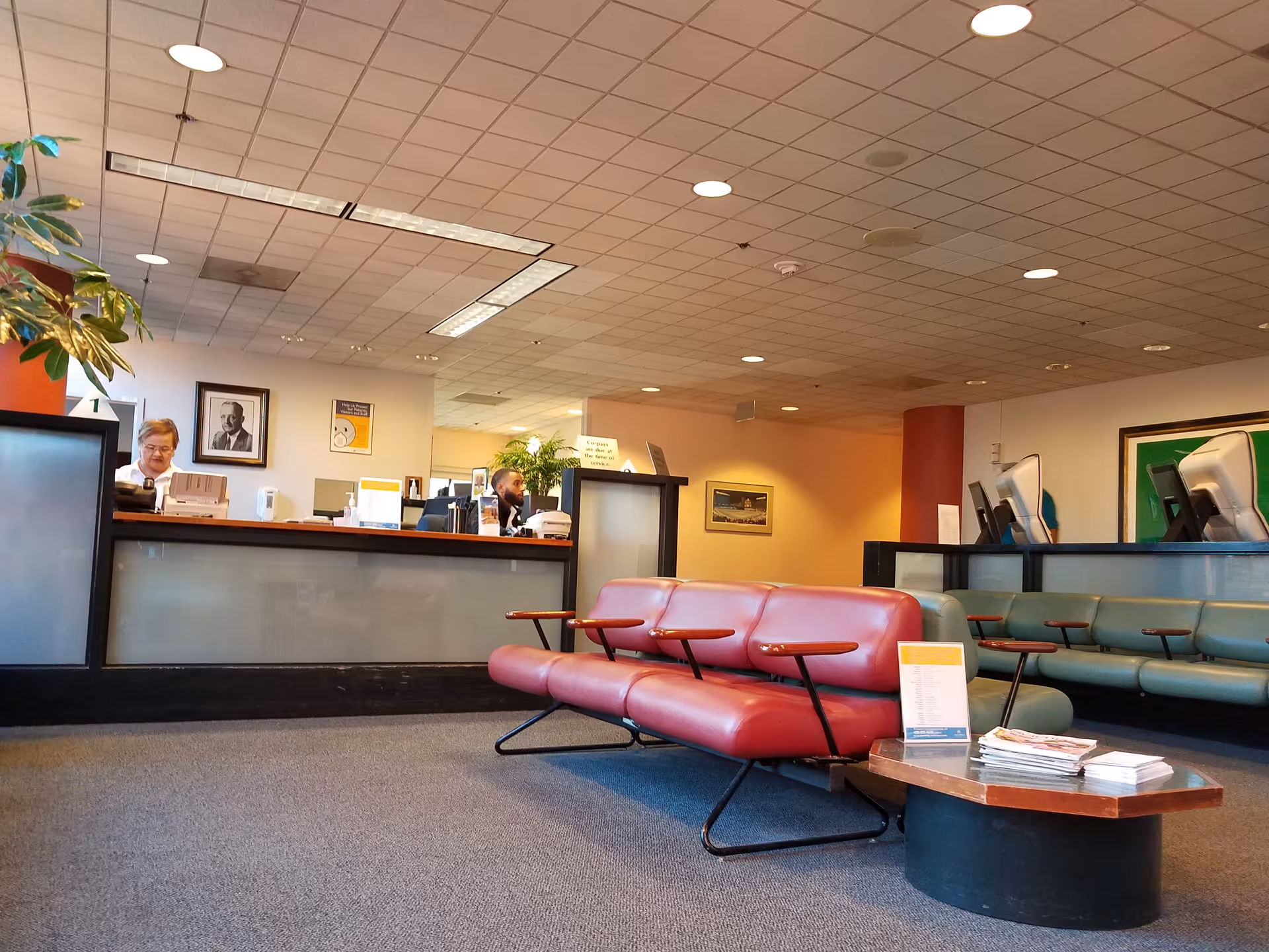 Hospital outpatient lobby with a red sofa, teal seating, front desk, and staff at computers.