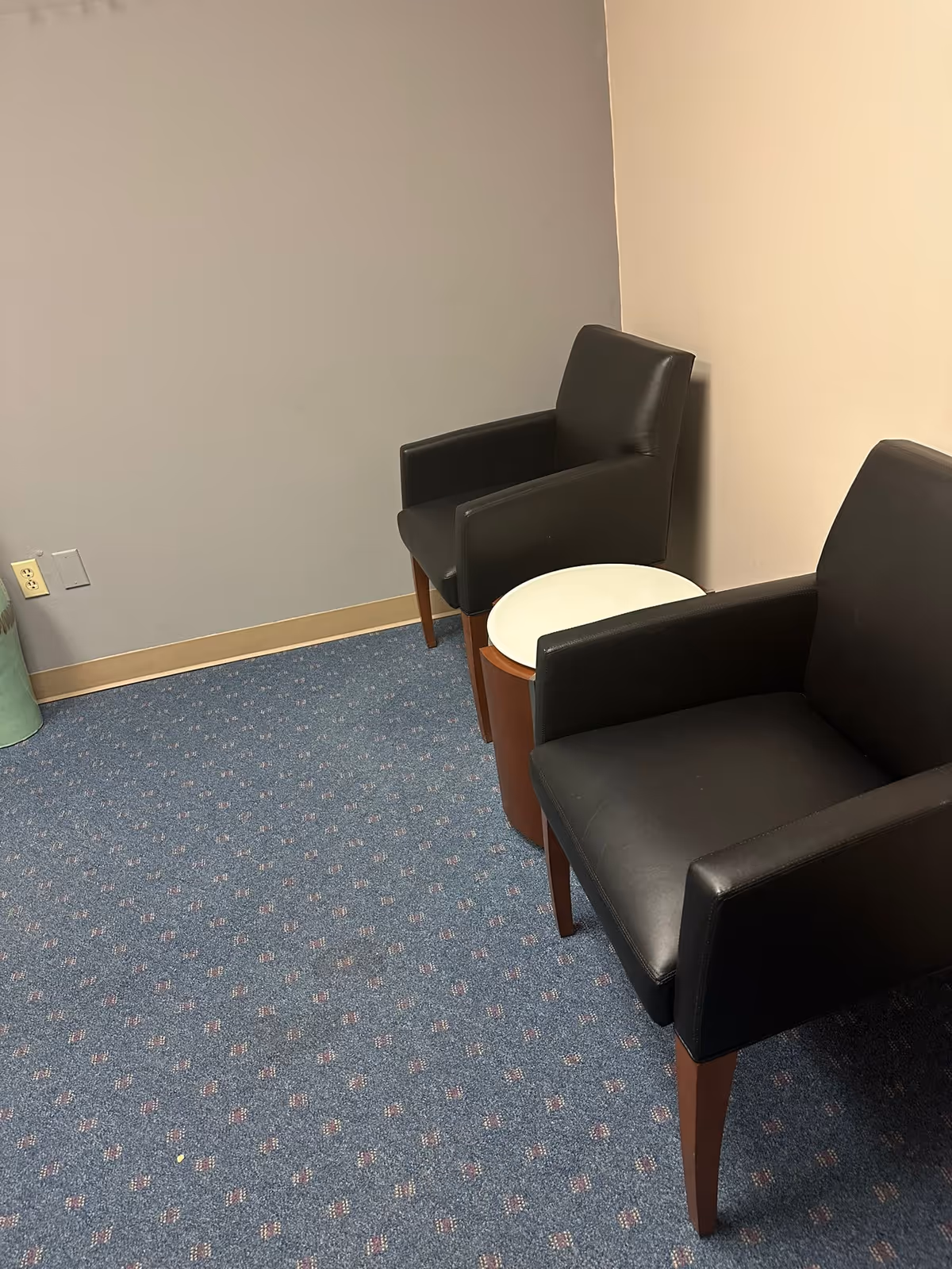 Two black leather armchairs flank a small round table in a blue-carpeted waiting area.
