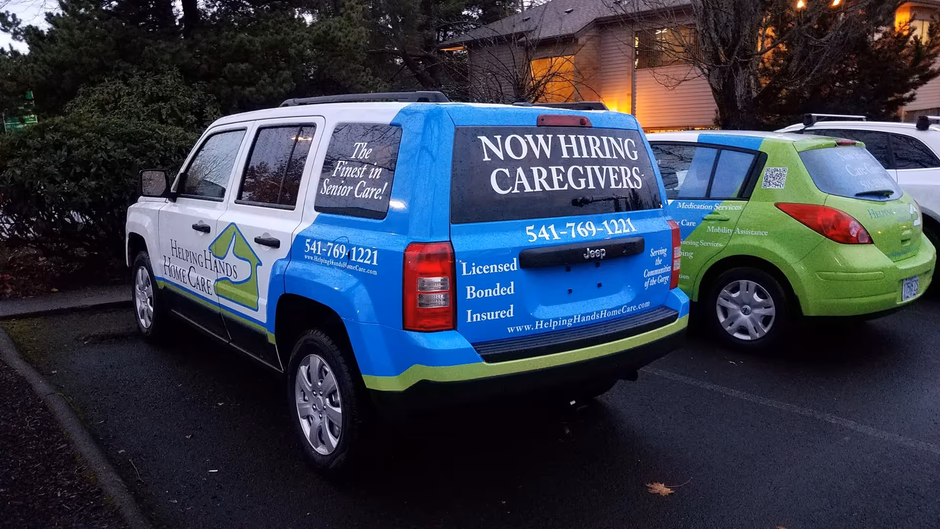Blue and green caregiving vans parked outside a building with caregiver recruitment signs.