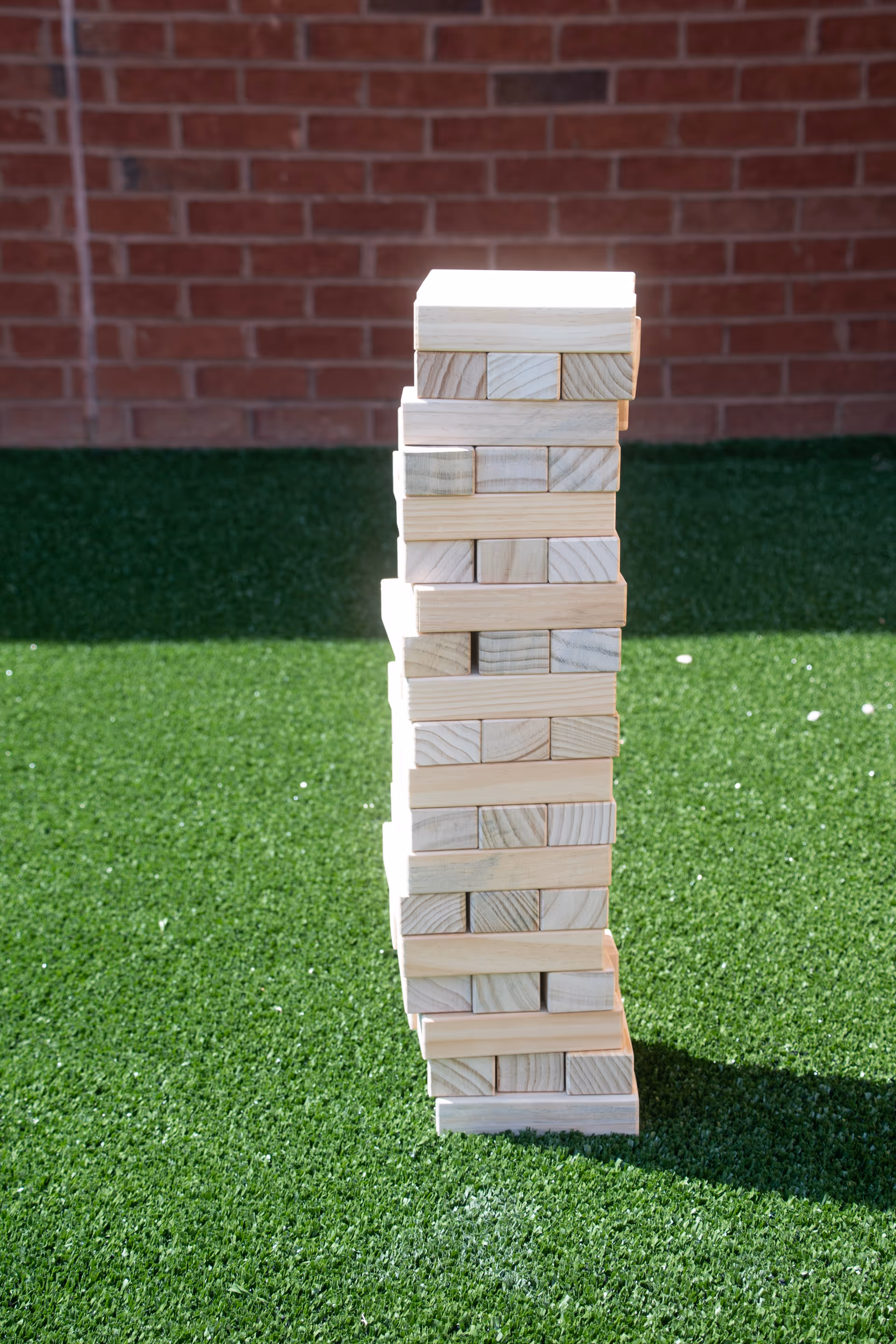 A tall wooden Jenga tower stands on bright green artificial grass outdoors.