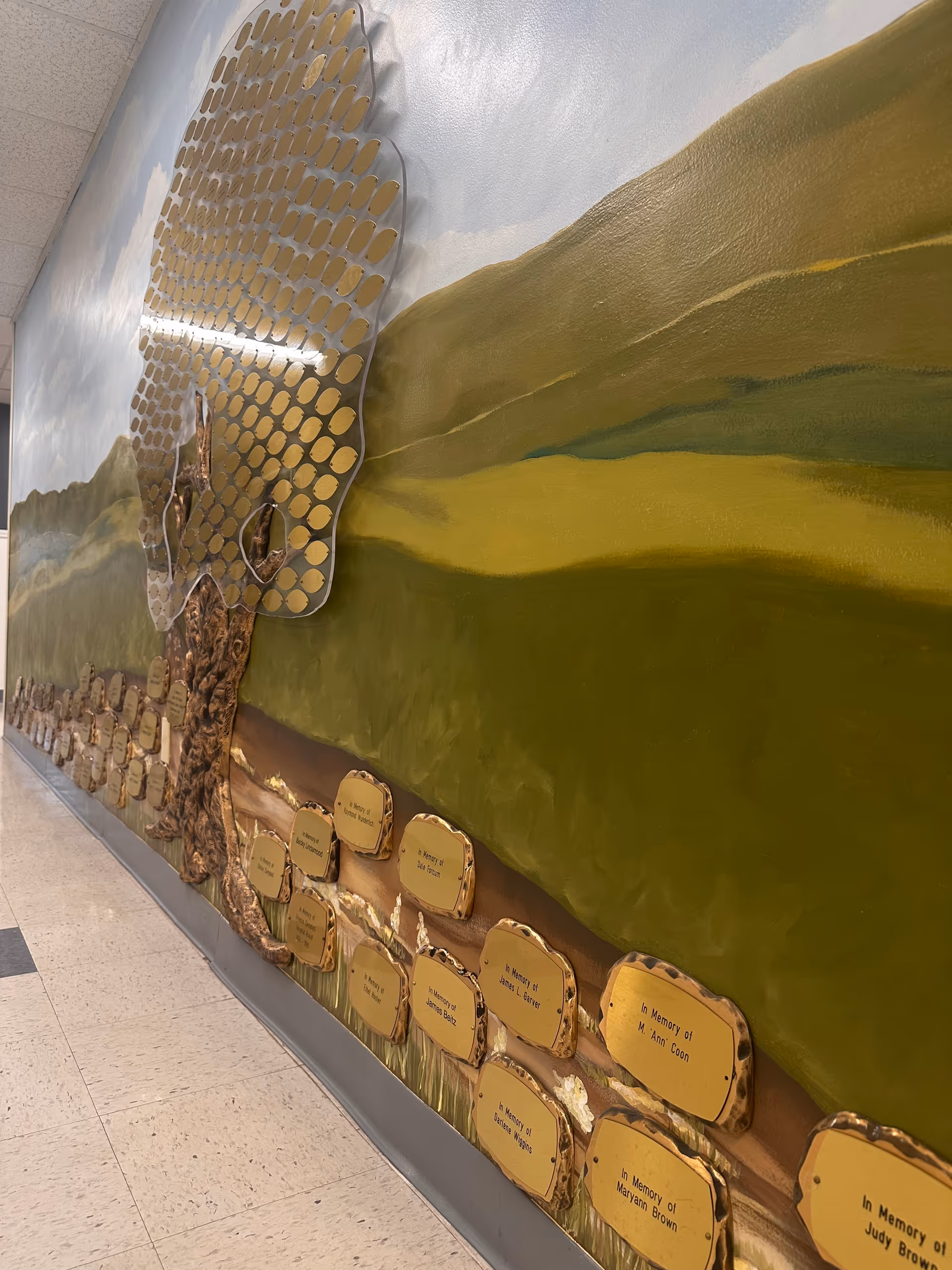 Indoor hallway featuring a large mural tree and gold memorial plaques along the wall.
