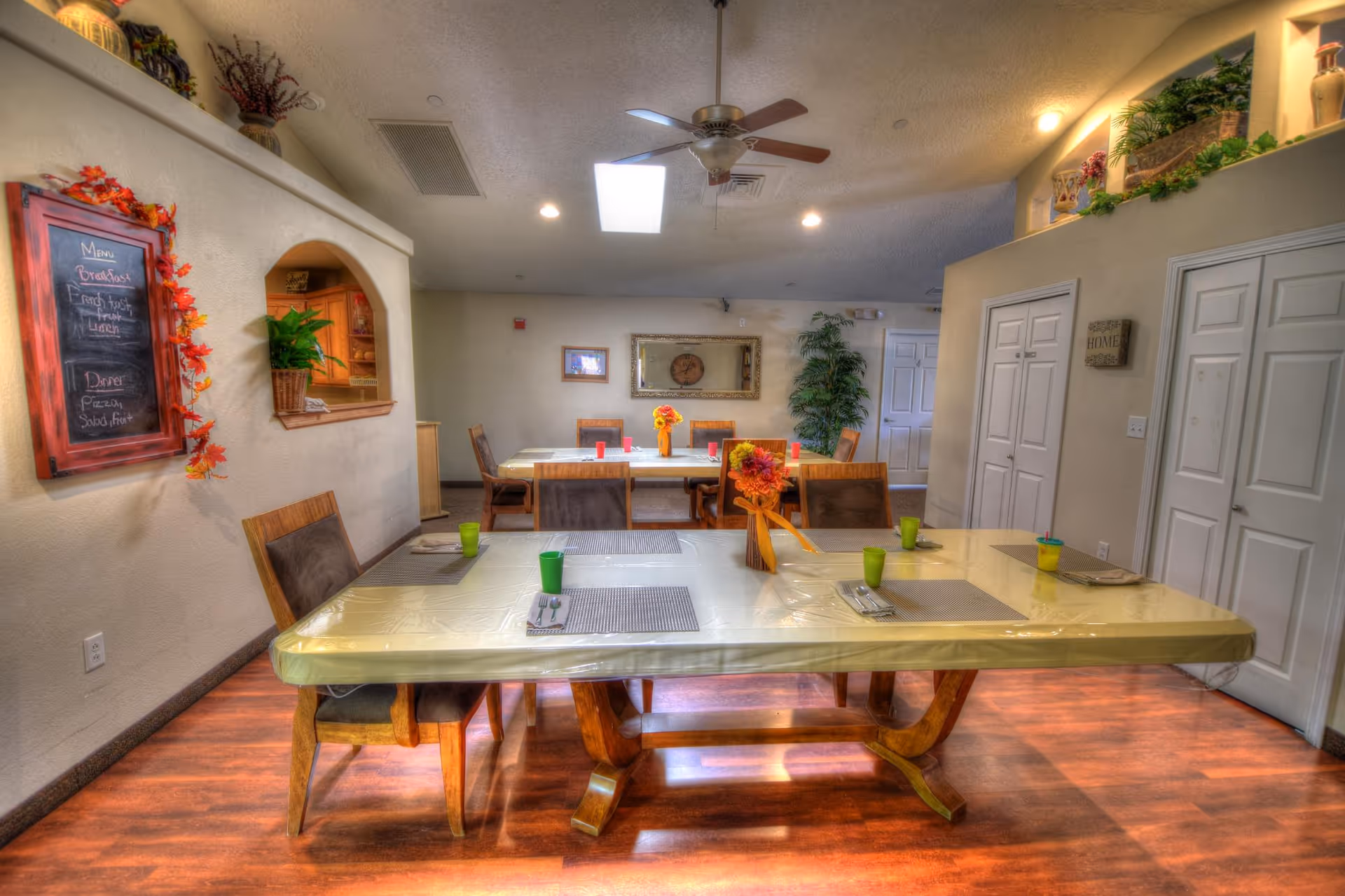 Sunlit dining room with long plastic-covered tables, chairs, and autumn decor in a senior living center.