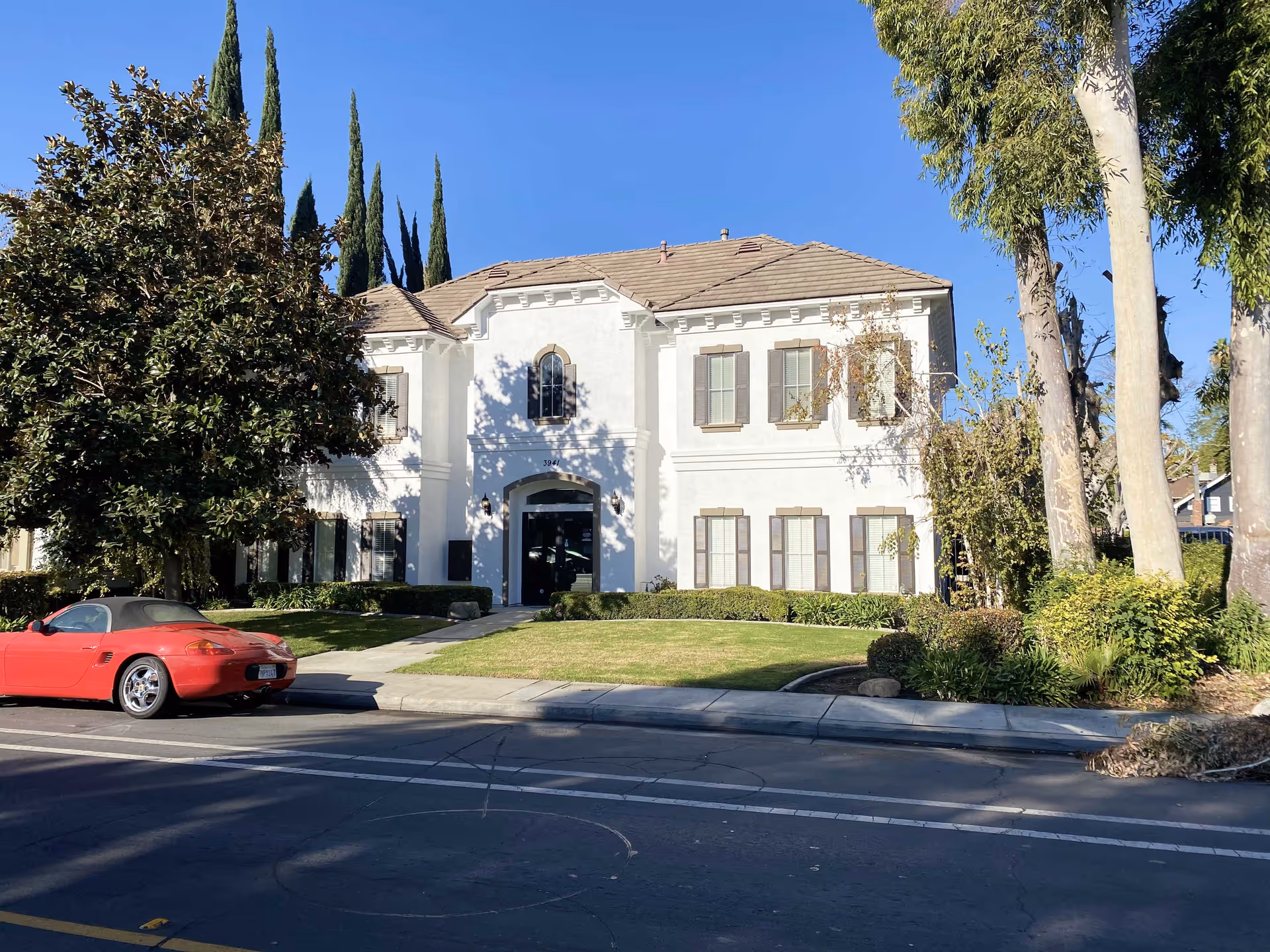 White two-story building with arched entrance, manicured lawn, red sports car, and trees.