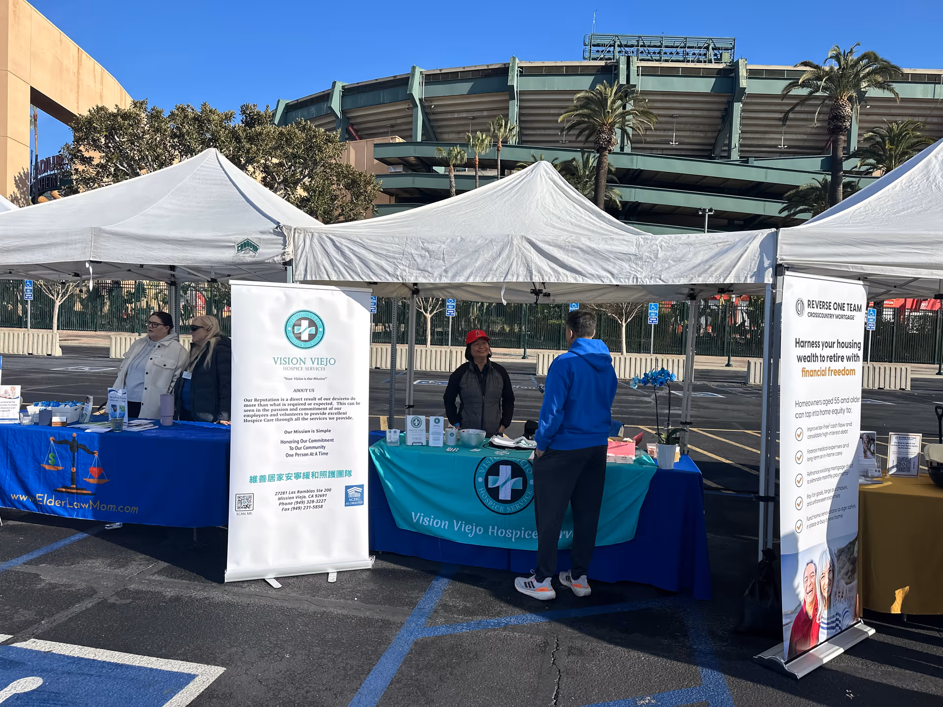 Outdoor charity fair with Vision Viejo Hospice banners under white canopies and tables.