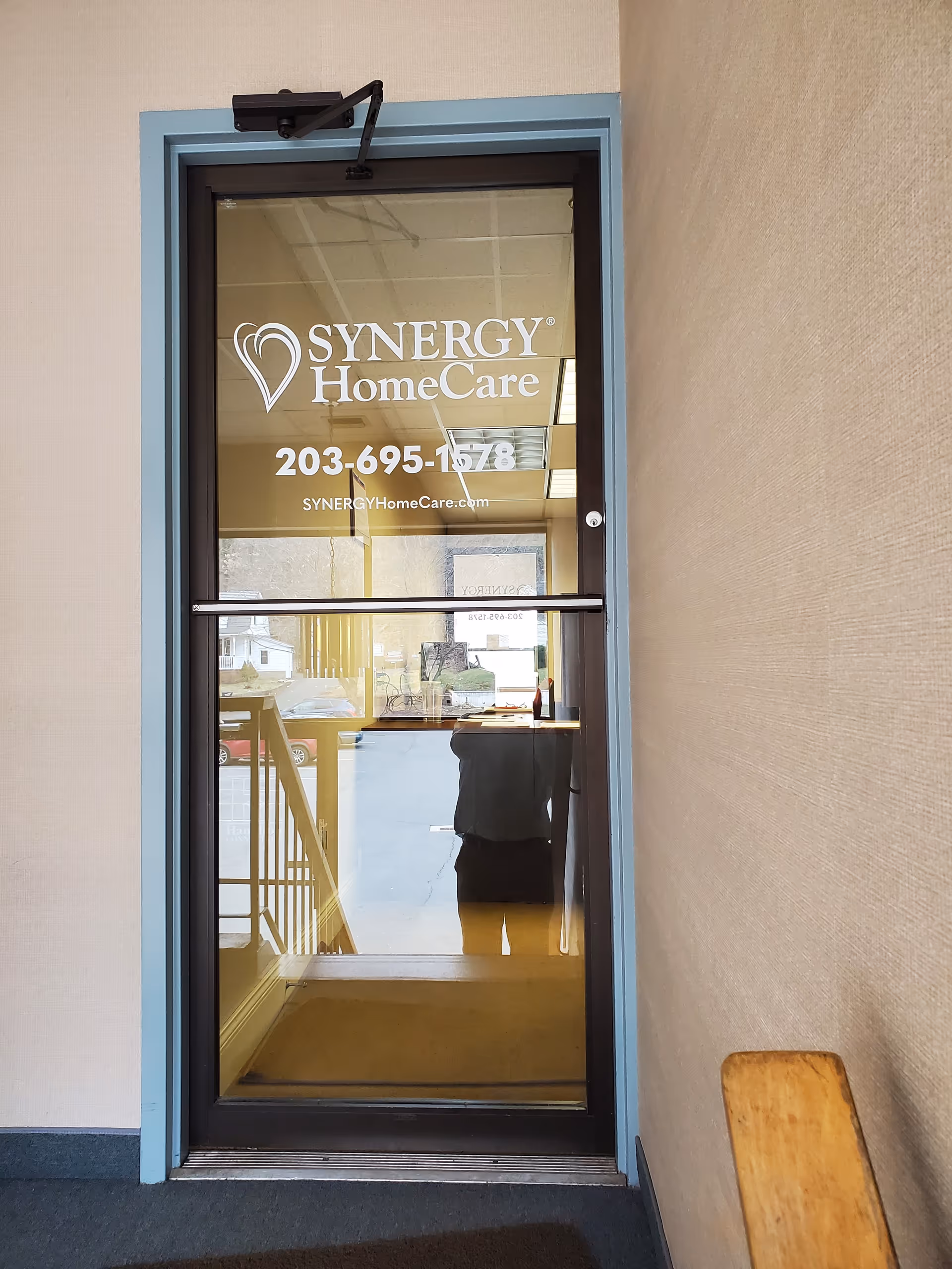 Glass entry door displays SYNERGY HomeCare logo and contact number inside a hallway.