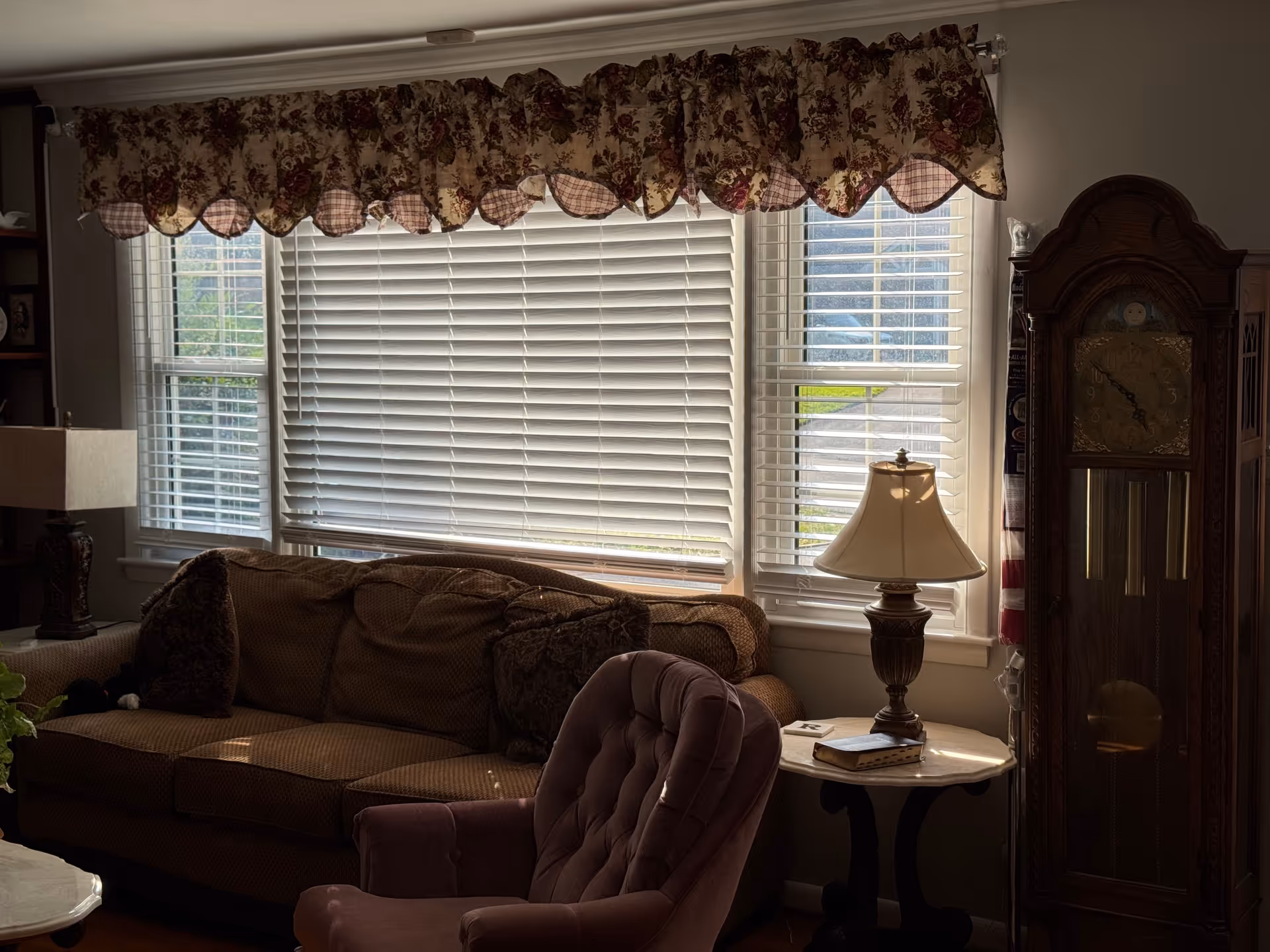Cozy living room with window blinds, sofa, armchair, side table, lamp, and grandfather clock.
