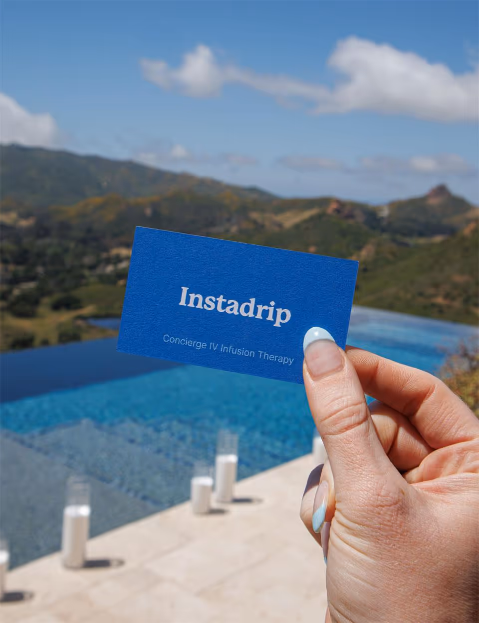 A hand holds a blue Instadrip card above a pool with mountains in background.