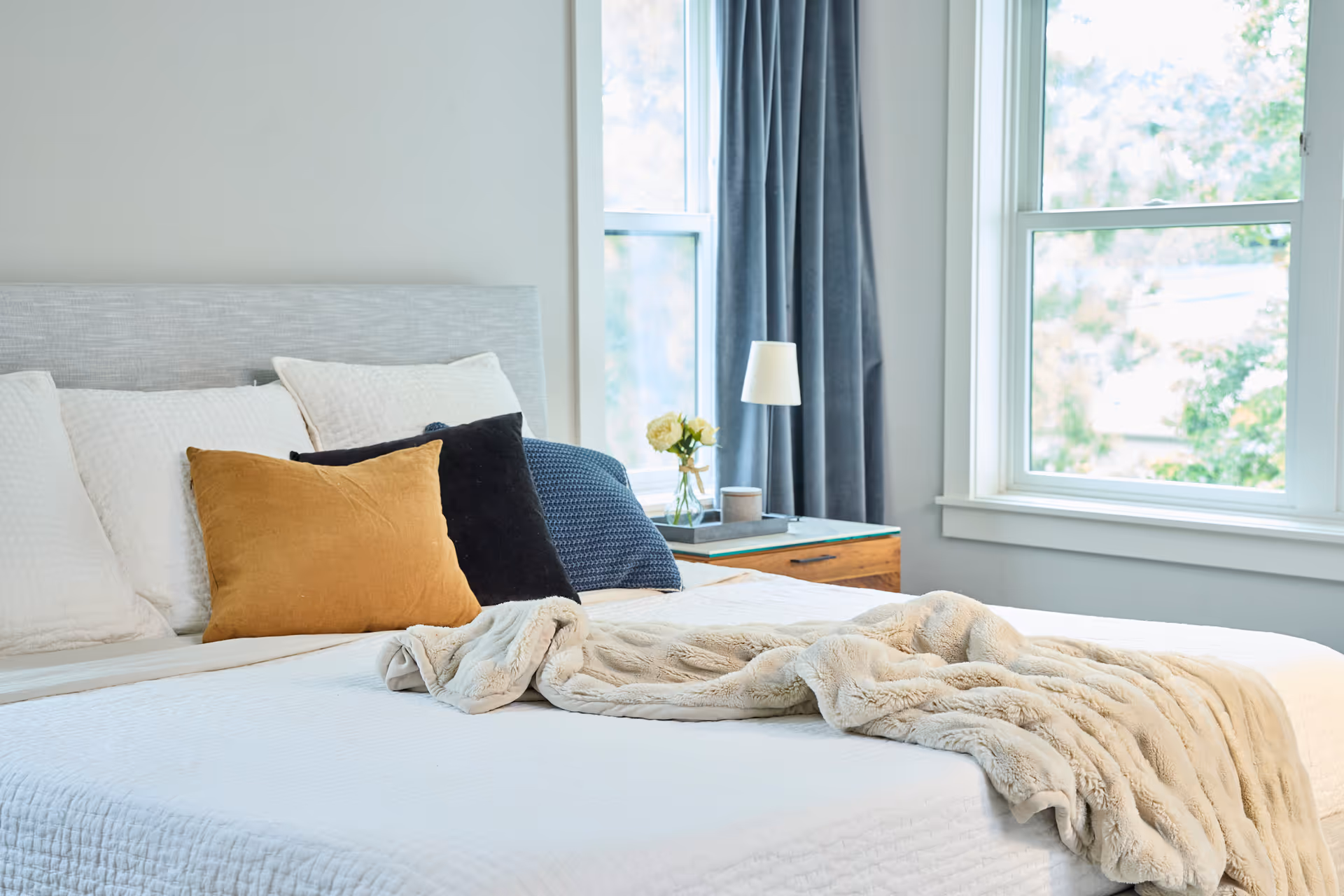 A neatly made bed in a bright bedroom with pillows, blanket, and nightstand.