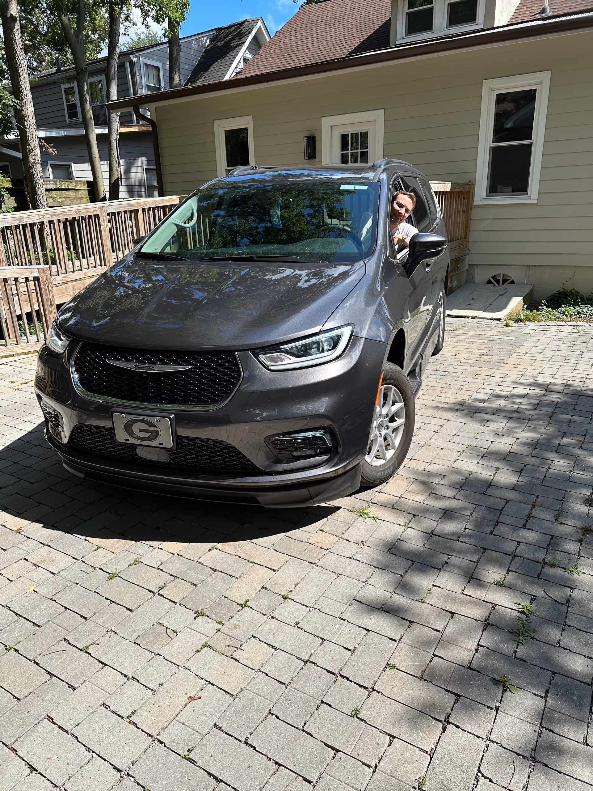 A gray SUV in a brick driveway beside a beige house, with a person inside.
