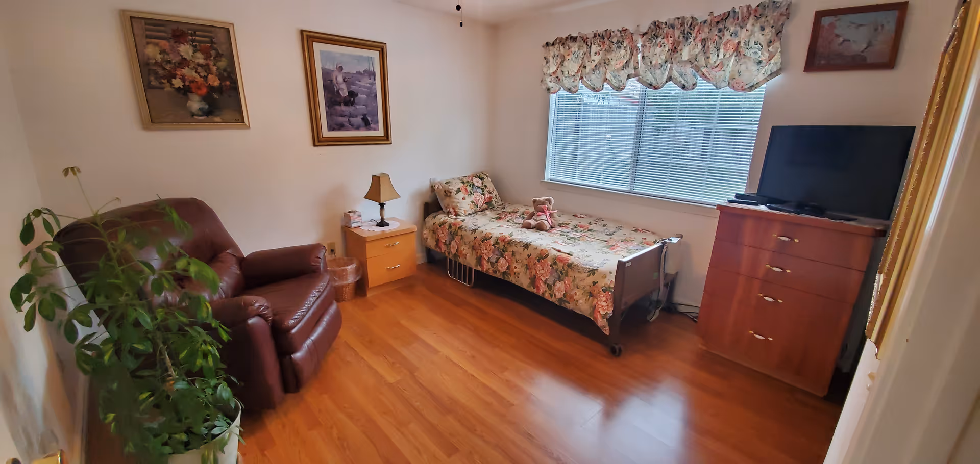 Cozy bedroom in a senior care facility with a floral bedspread, bed, dresser, TV, and seating.