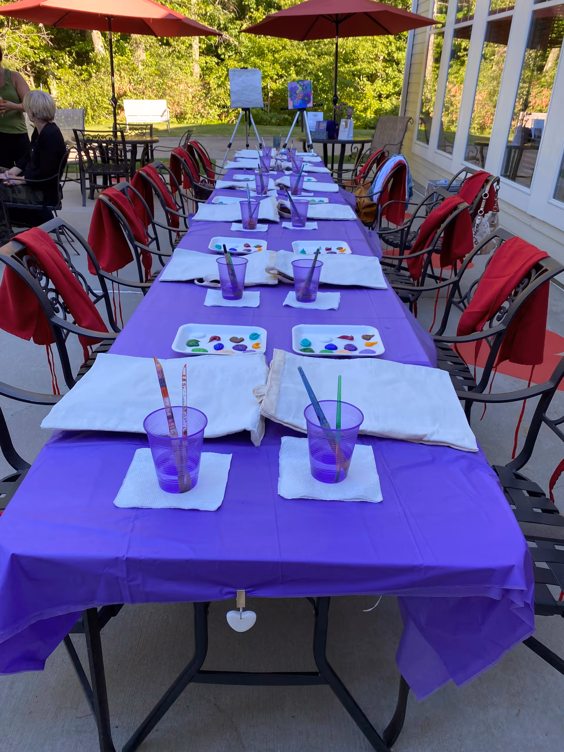 Long outdoor table with purple tablecloth, paint plates, cups, and napkins under red umbrellas.