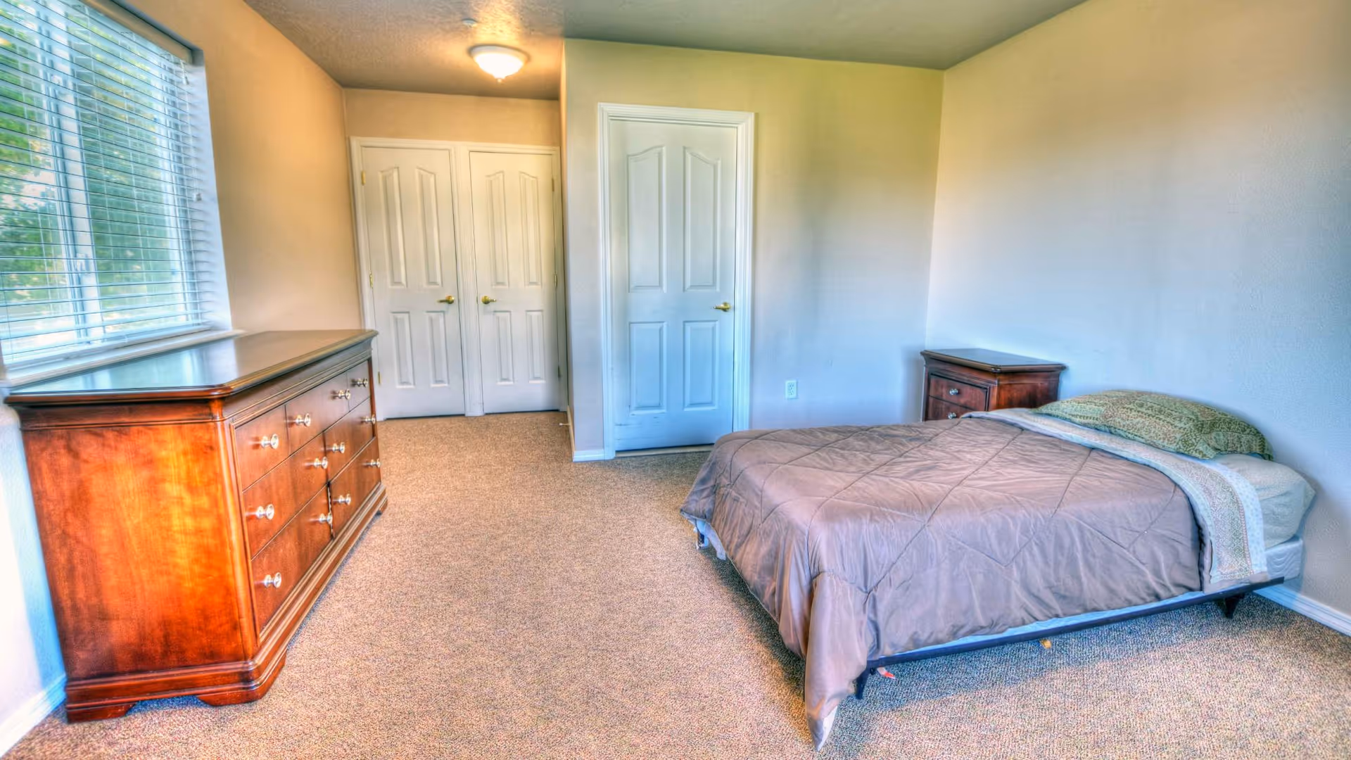 A furnished bedroom with a bed, dresser, window, and closet doors in a plain room.