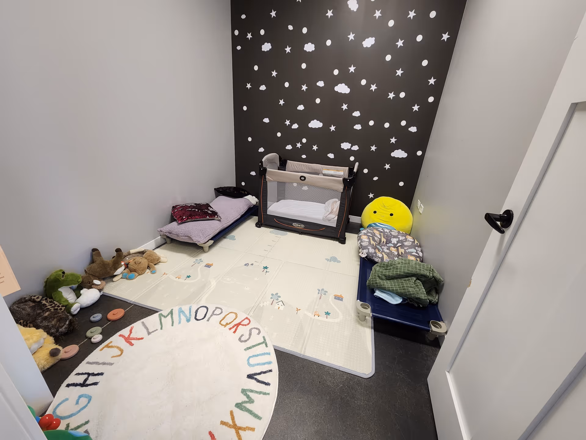 Small children's play area with a playpen, soft toys, and alphabet rug in a bright room.