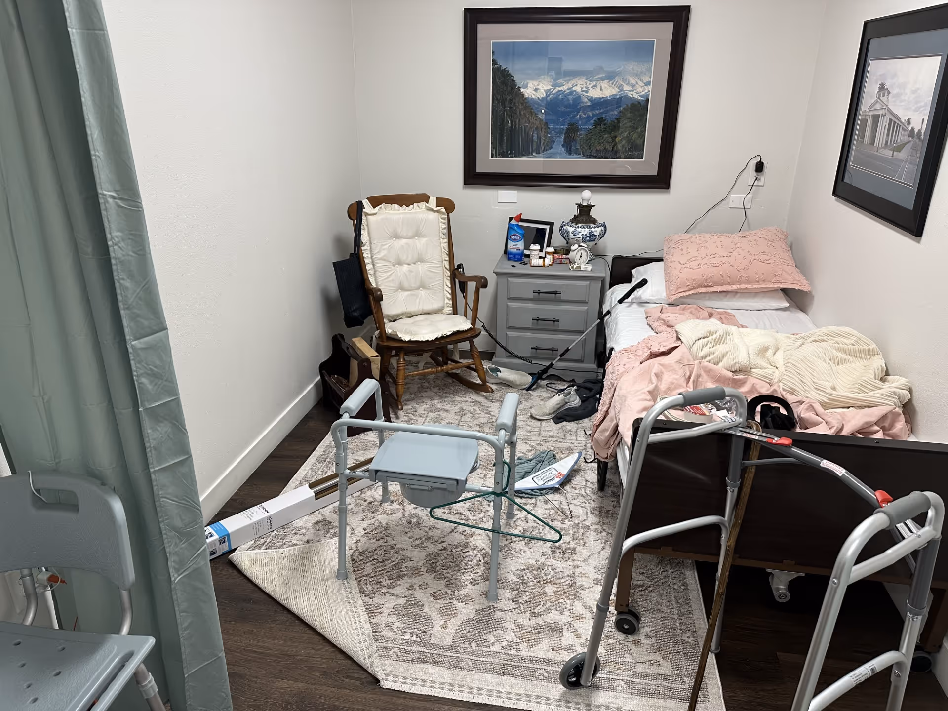 Small patient bedroom with bed, walker, chair, nightstand, blankets, and framed pictures.