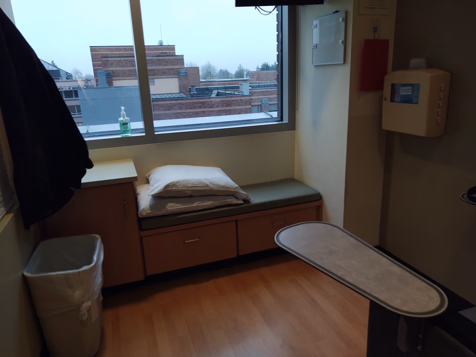 Interior hospital-style patient room with bed, window seat, cabinet, and movable table.
