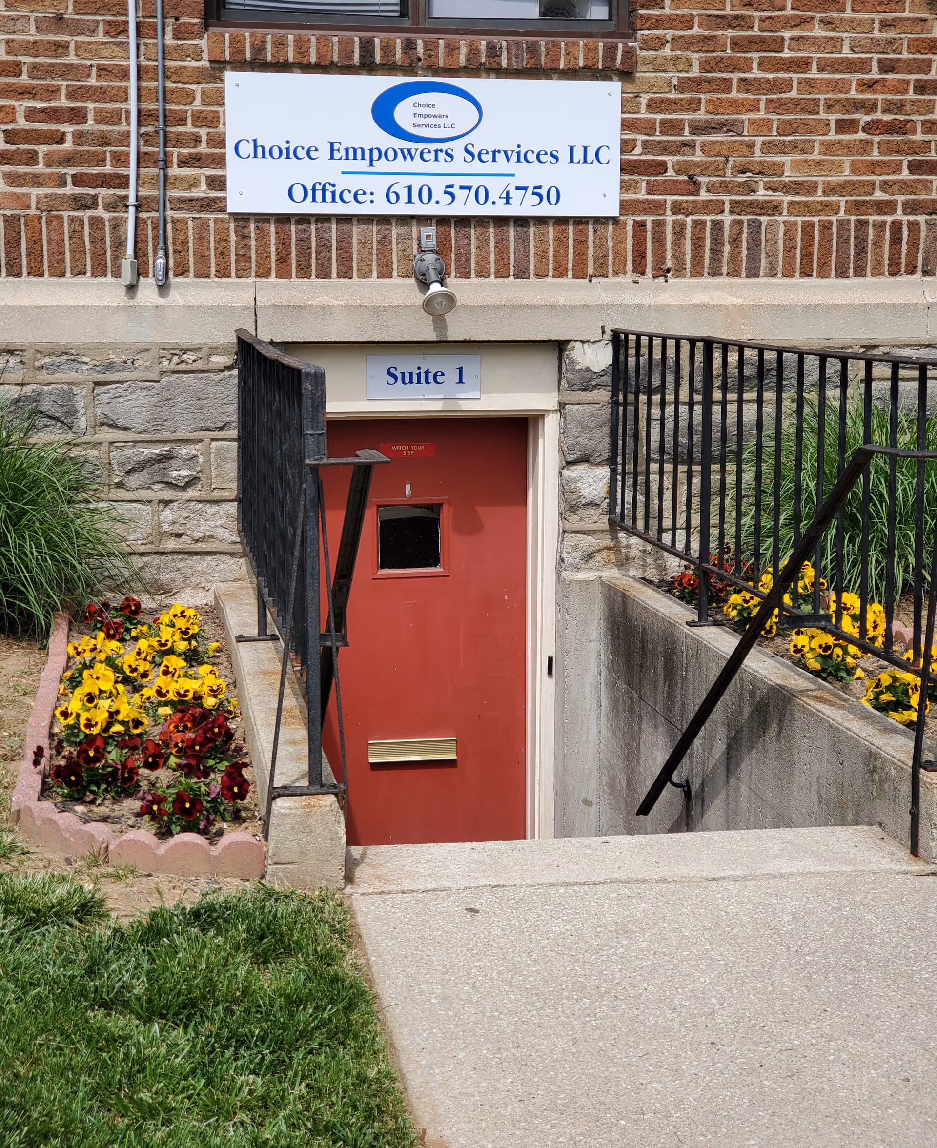 Exterior brick building entrance with a red basement door, flowers, and a Choice Empowers Services sign.