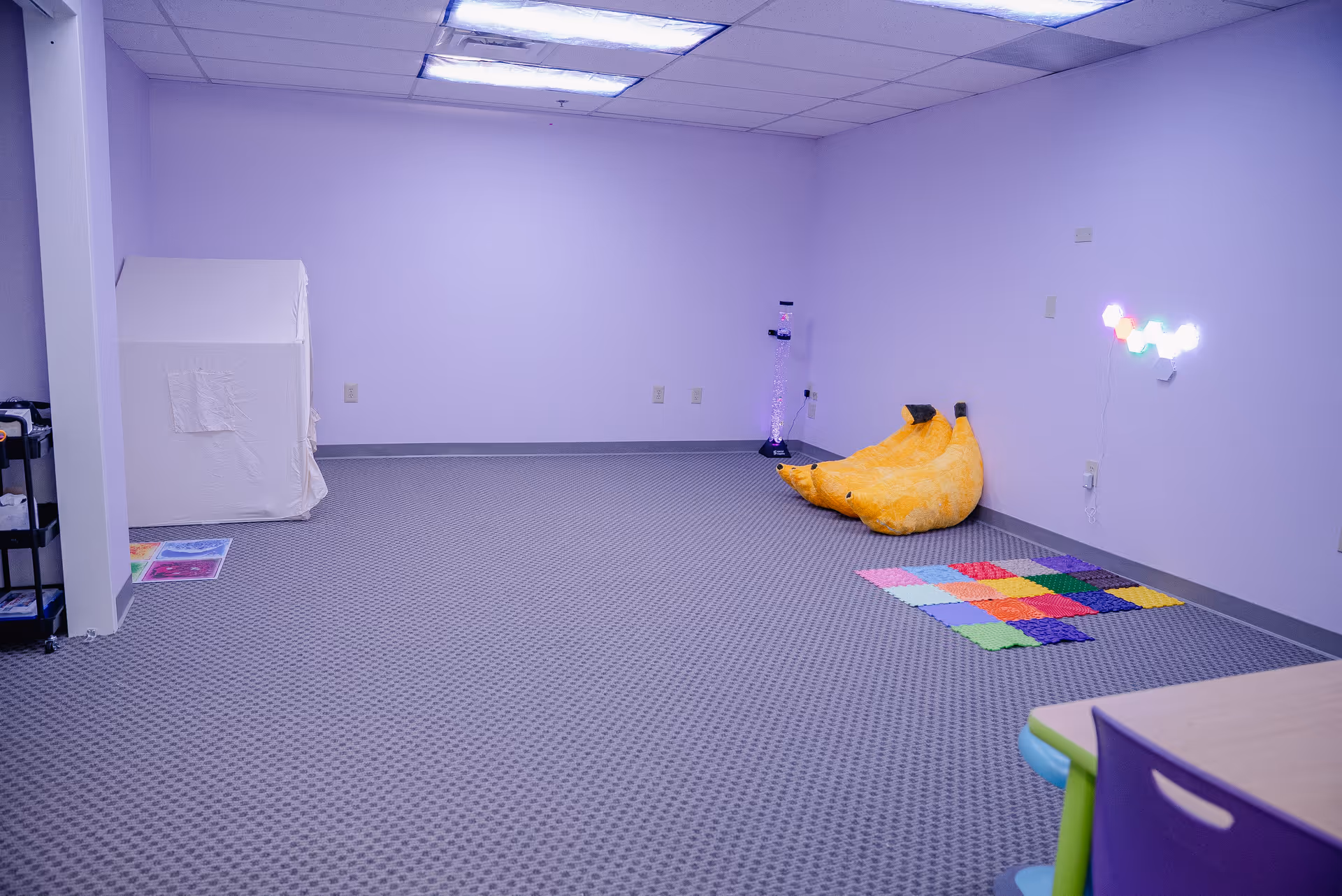 Pastel therapy playroom with mats, banana bean bag, neon lights, and table.