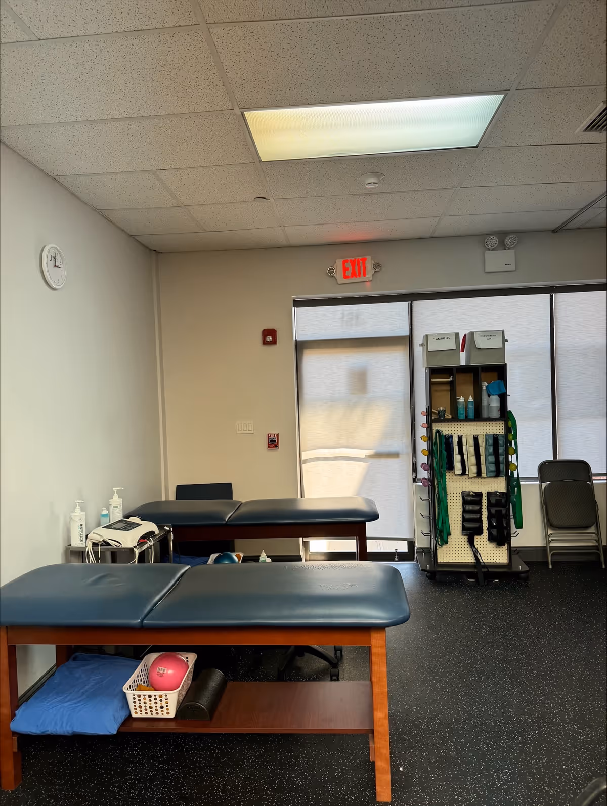 A physical therapy treatment room with two blue padded tables and equipment shelves.