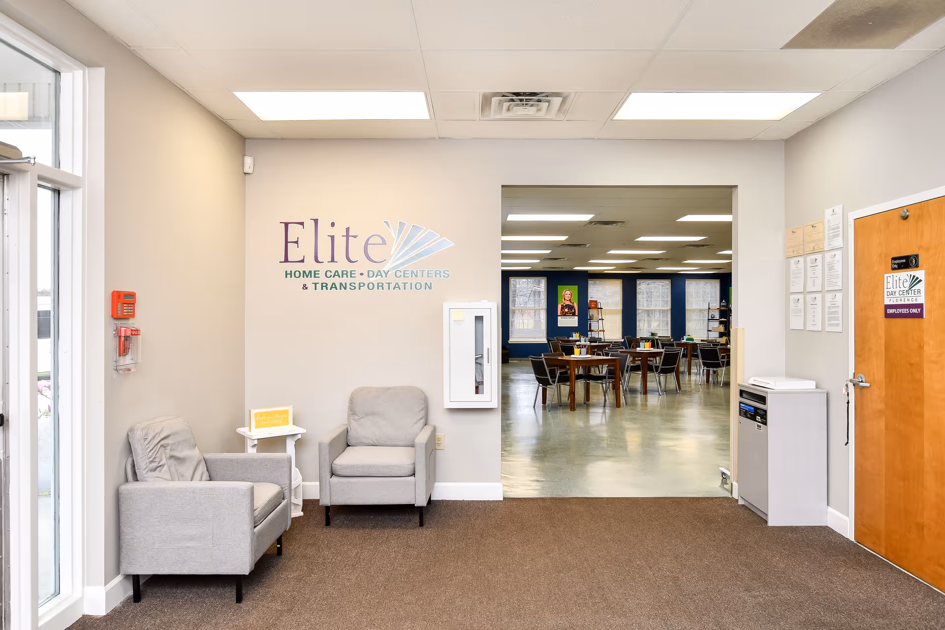 Interior lobby area with two gray chairs, logo on wall, and doorway to a dining room.