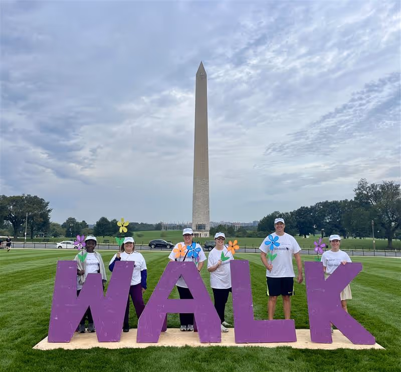 Group of people posing with colorful flower props behind large purple WALK letters near Washington Monument.
