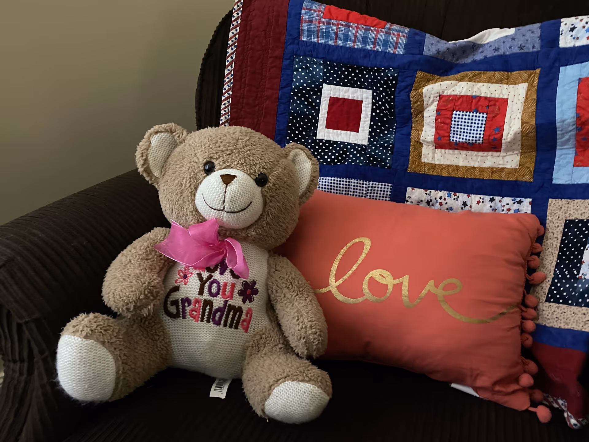 Teddy bear with pink bow sits on a couch beside a colorful quilt and heart pillow.