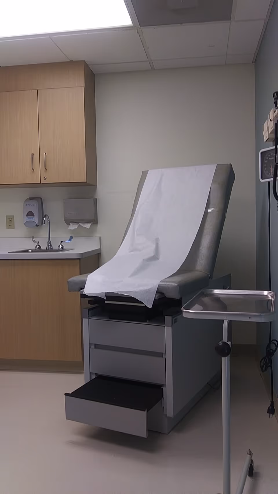 Hospital exam room with a paper-covered exam chair, sink and cabinets.