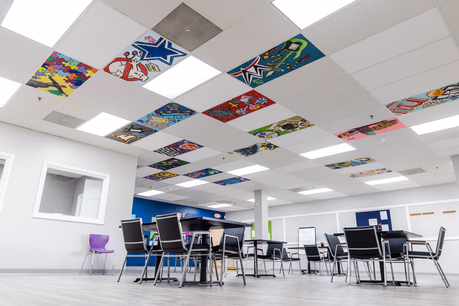 Bright communal dining area with several tables and black chairs beneath colorful ceiling art.