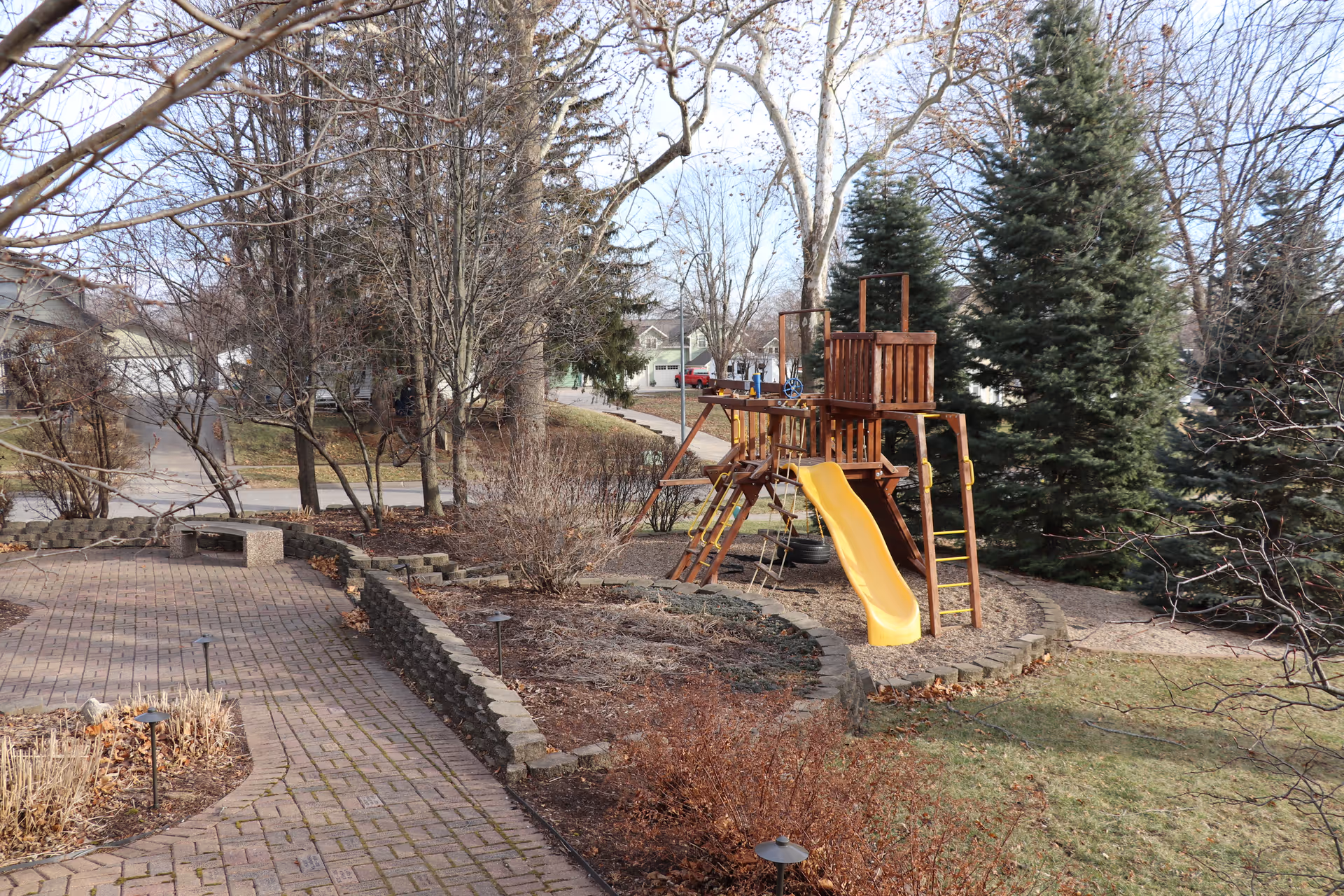 Outdoor courtyard featuring a wooden playset with a yellow slide, paving, and trees.