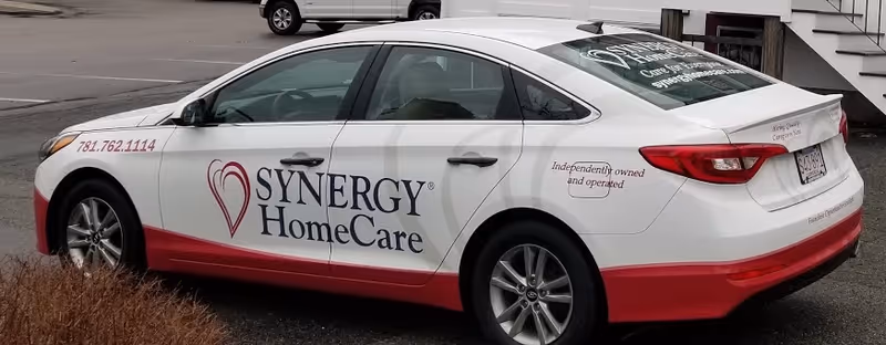 White SYNERGY HomeCare promotional car parked in a lot, displaying logo and contact info.