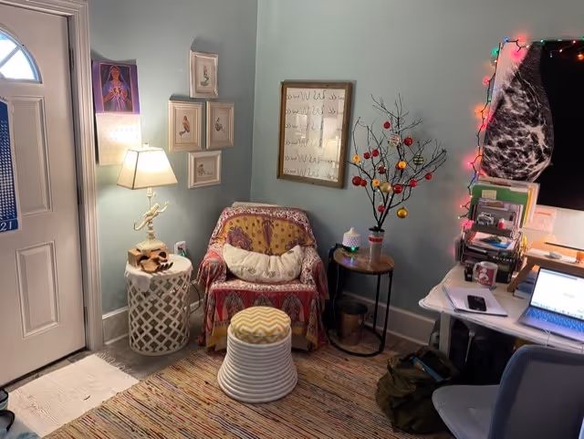 Cozy living corner near entrance with patterned chair, lamp, wall art, ornamental tree, and desk.