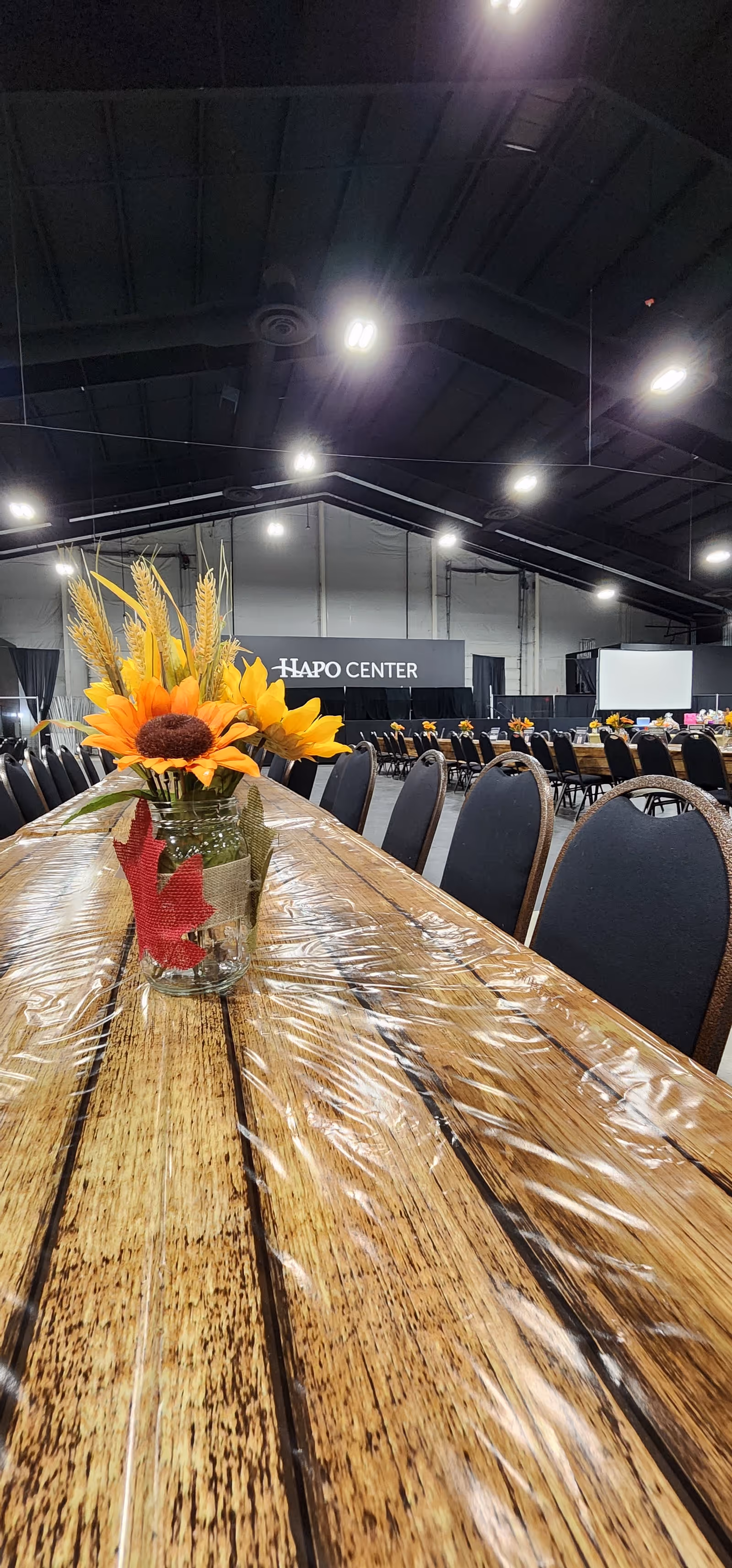 Long wooden table with sunflowers centerpiece in a large, empty indoor dining hall.