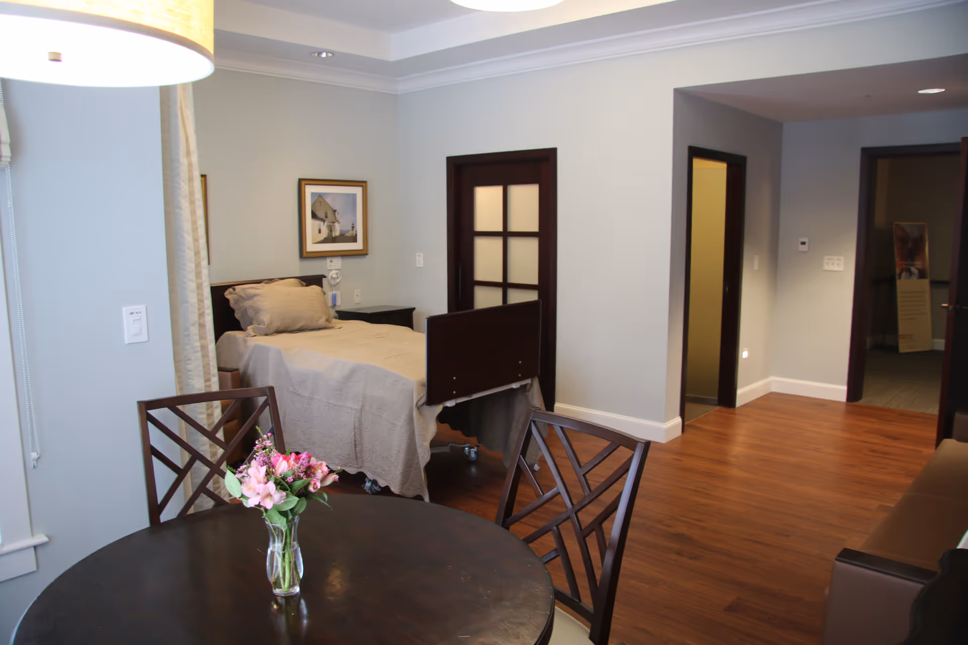 Interior of a senior living suite with a bed, round dining table, and flowers.