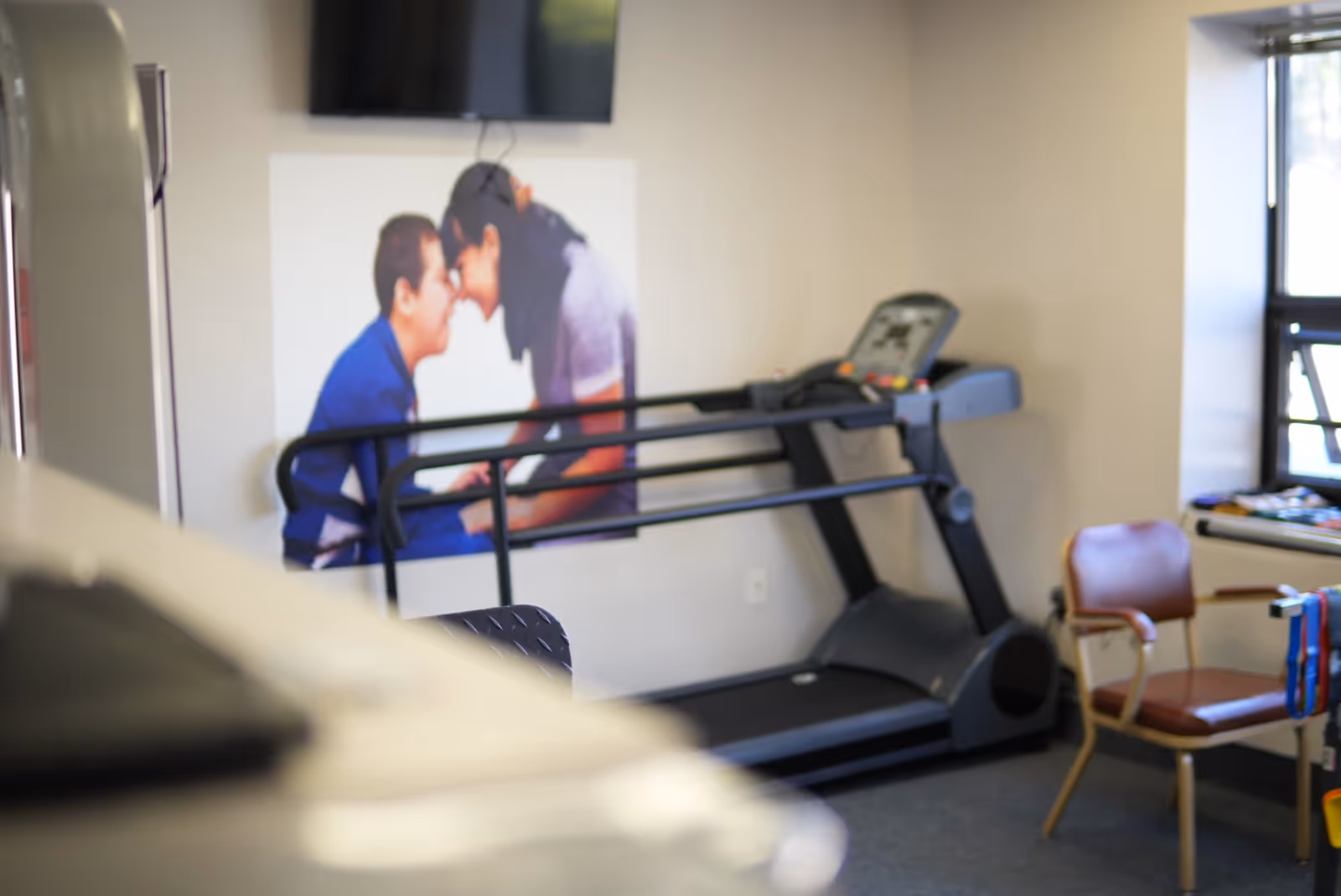 A treadmill and exercise area inside a senior living facility, with a chair and wall art.