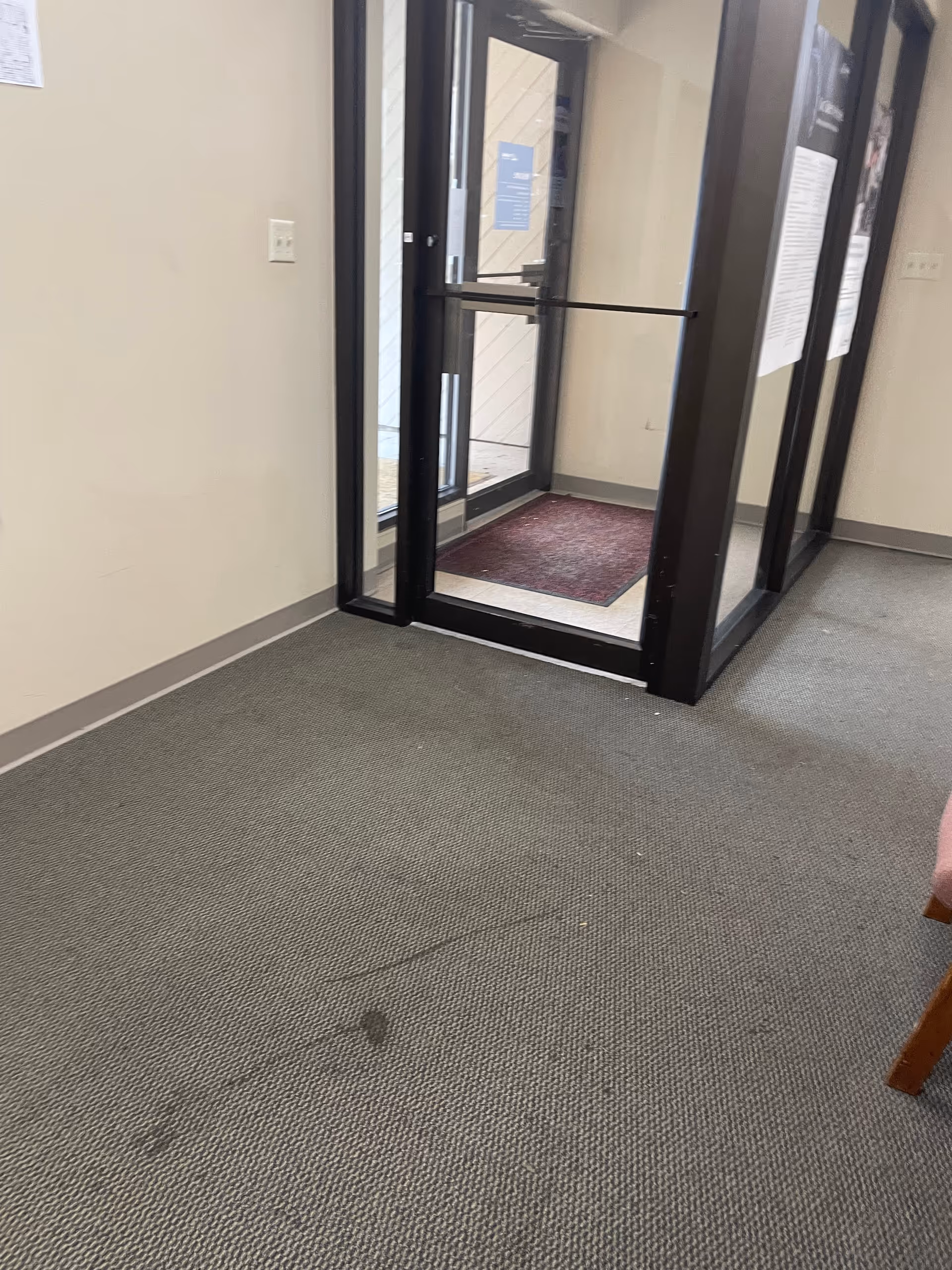 Interior entrance area with glass doors and a maroon mat in a neutral hallway.
