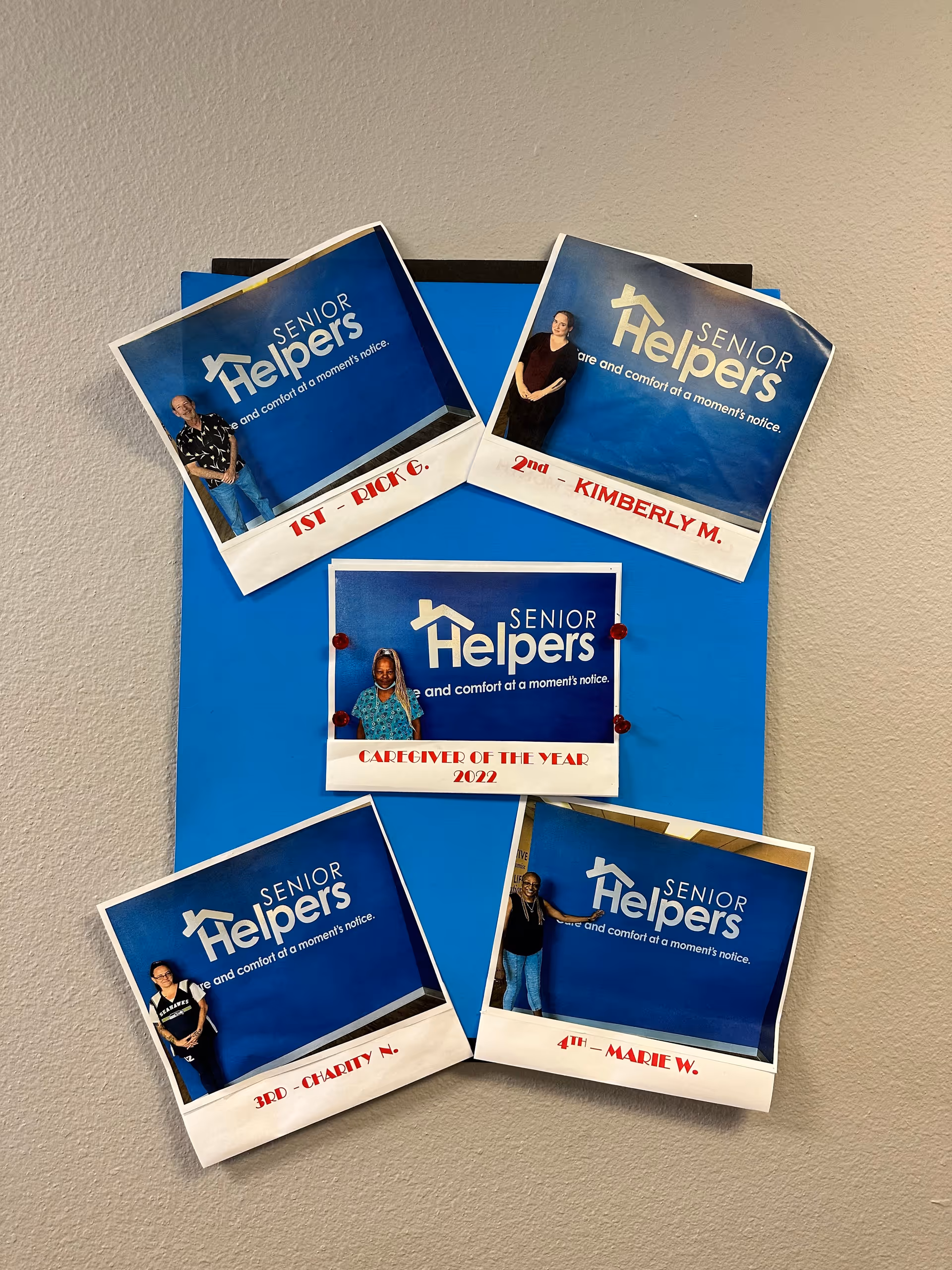 Bulletin board with Senior Helpers certificates and photos of caregivers on blue backdrop.