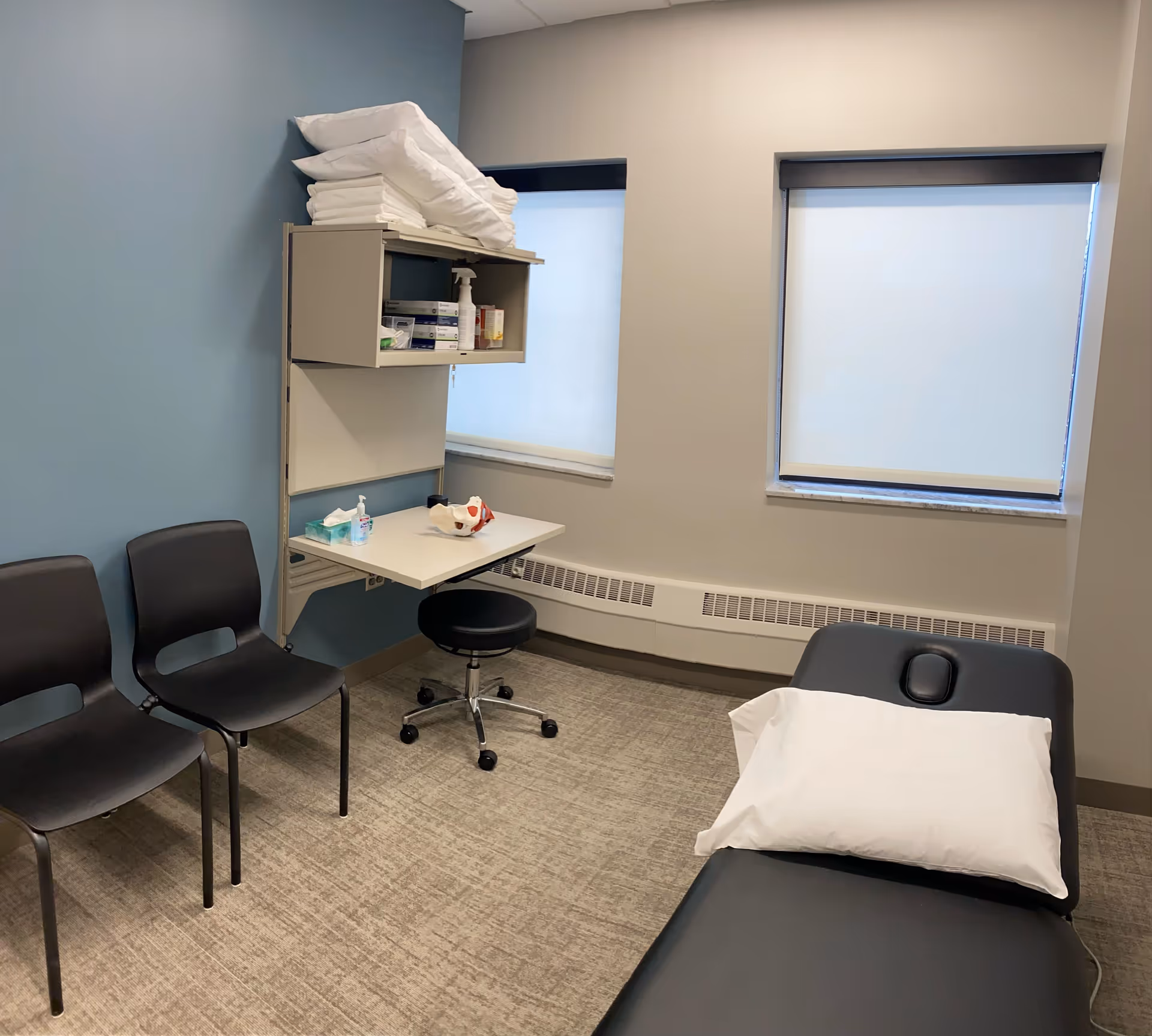 Medical examination room with an adjustable bed, stools, chairs, and shelves of supplies.