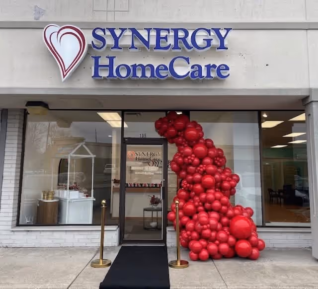 Front of SYNERGY HomeCare building with blue sign and red balloon arch at entrance.