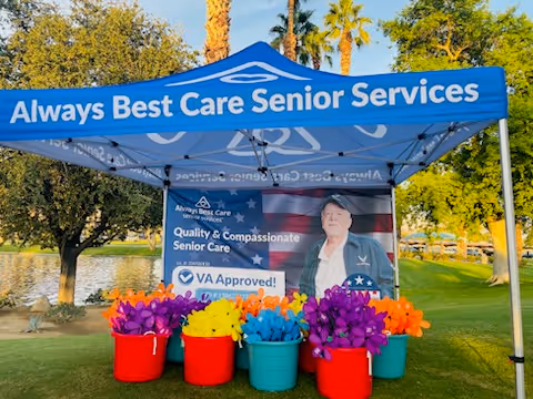 Outdoor blue canopy displays Always Best Care Senior Services beside colorful flower buckets.