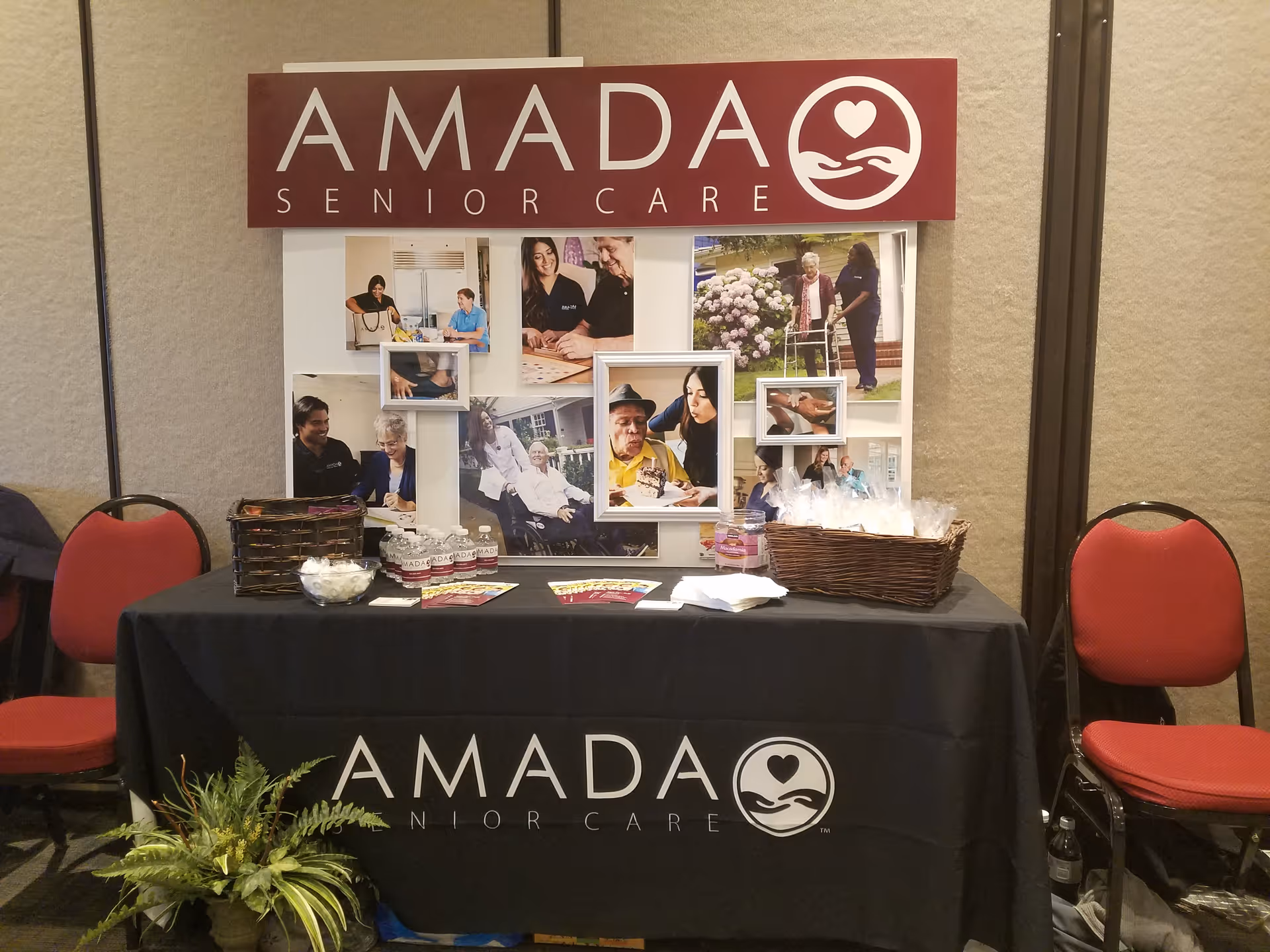 AMADA Senior Care display table with photos, banners, and chairs in an indoor event setup.