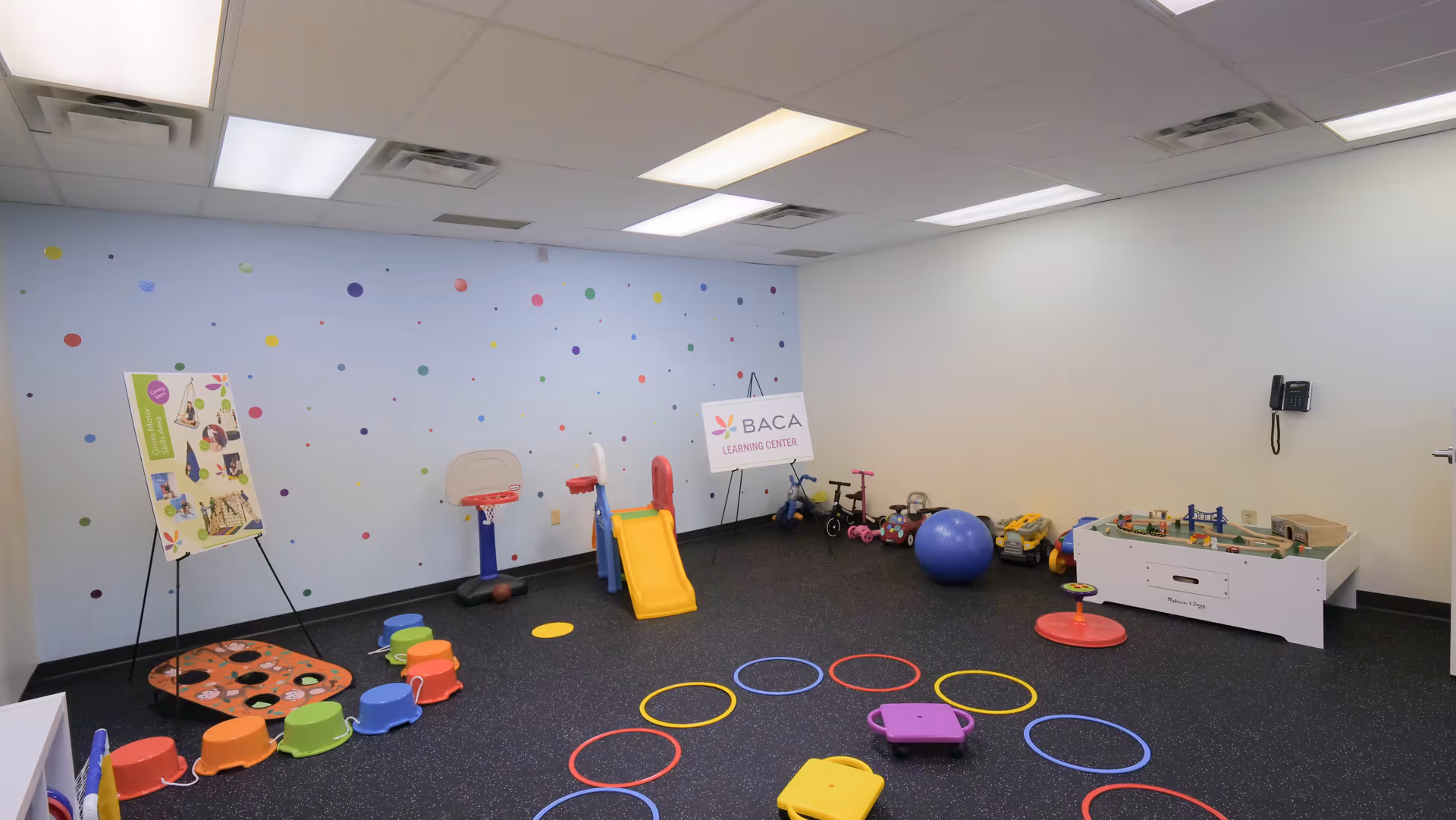 Bright indoor playroom with colorful therapy equipment, rings, slide, and polka-dot wall.