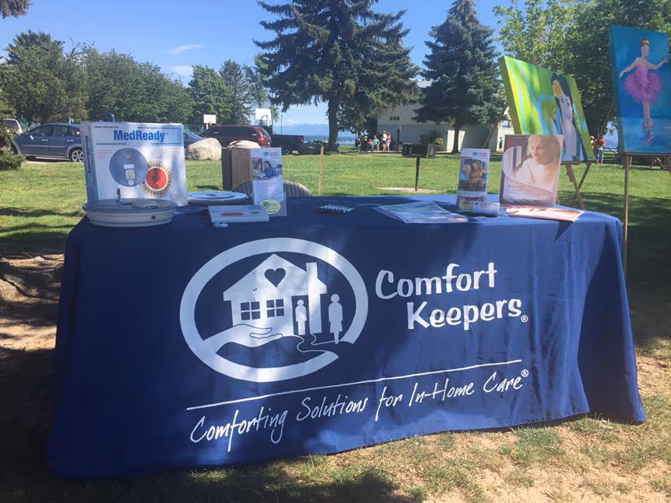 Outdoor event table draped with Comfort Keepers banner and promotional materials in a park.
