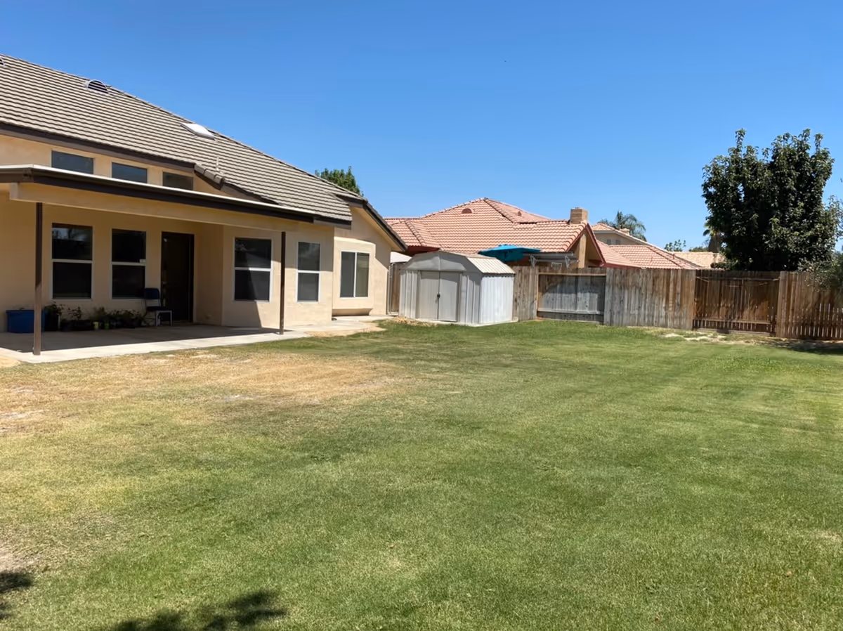 Backyard of a single-story house with a covered patio, shed, wooden fence, and blue sky.