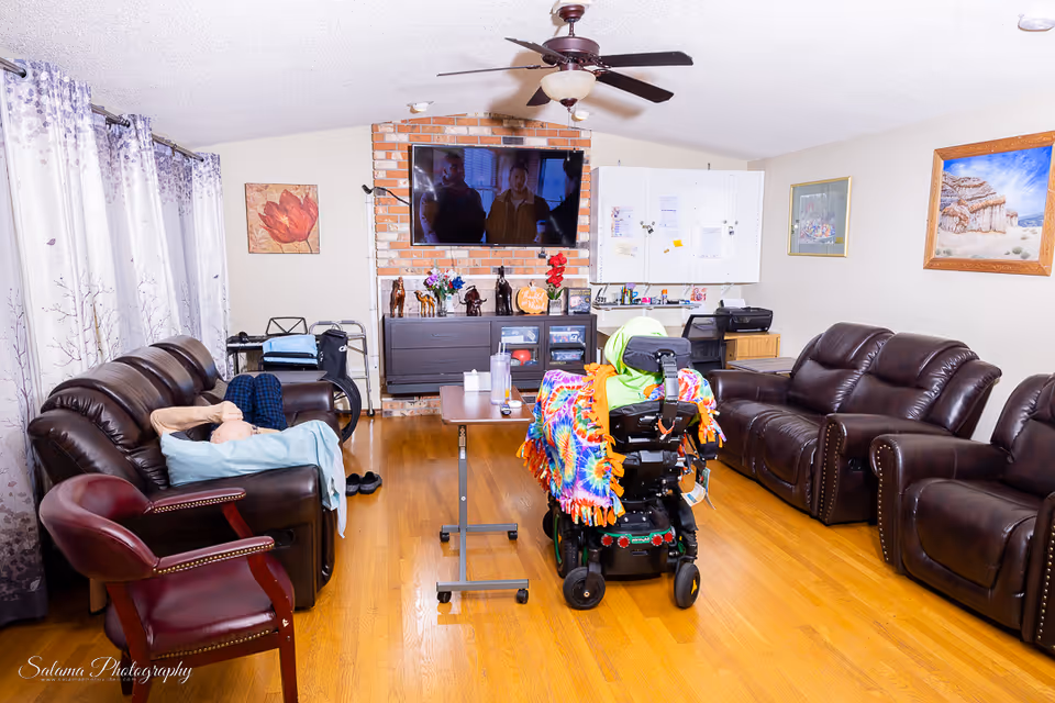 Cozy common living room with sofas, a recliner, wheelchair, TV, and brick fireplace.