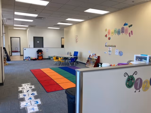 Bright open therapy room with colorful mats, small tables, chairs, and wall decorations.