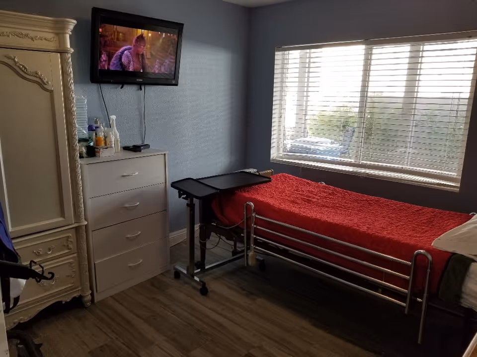 A small bedroom in a senior home with a bed, dresser, TV, and window blinds.