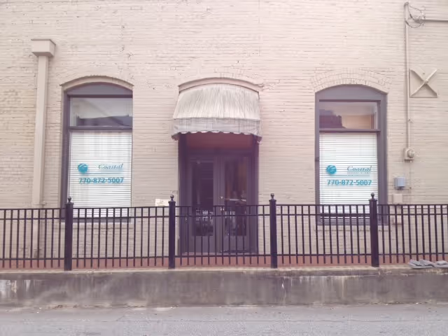 Exterior view of a brick building entrance with an awning, windows, and a black fence.