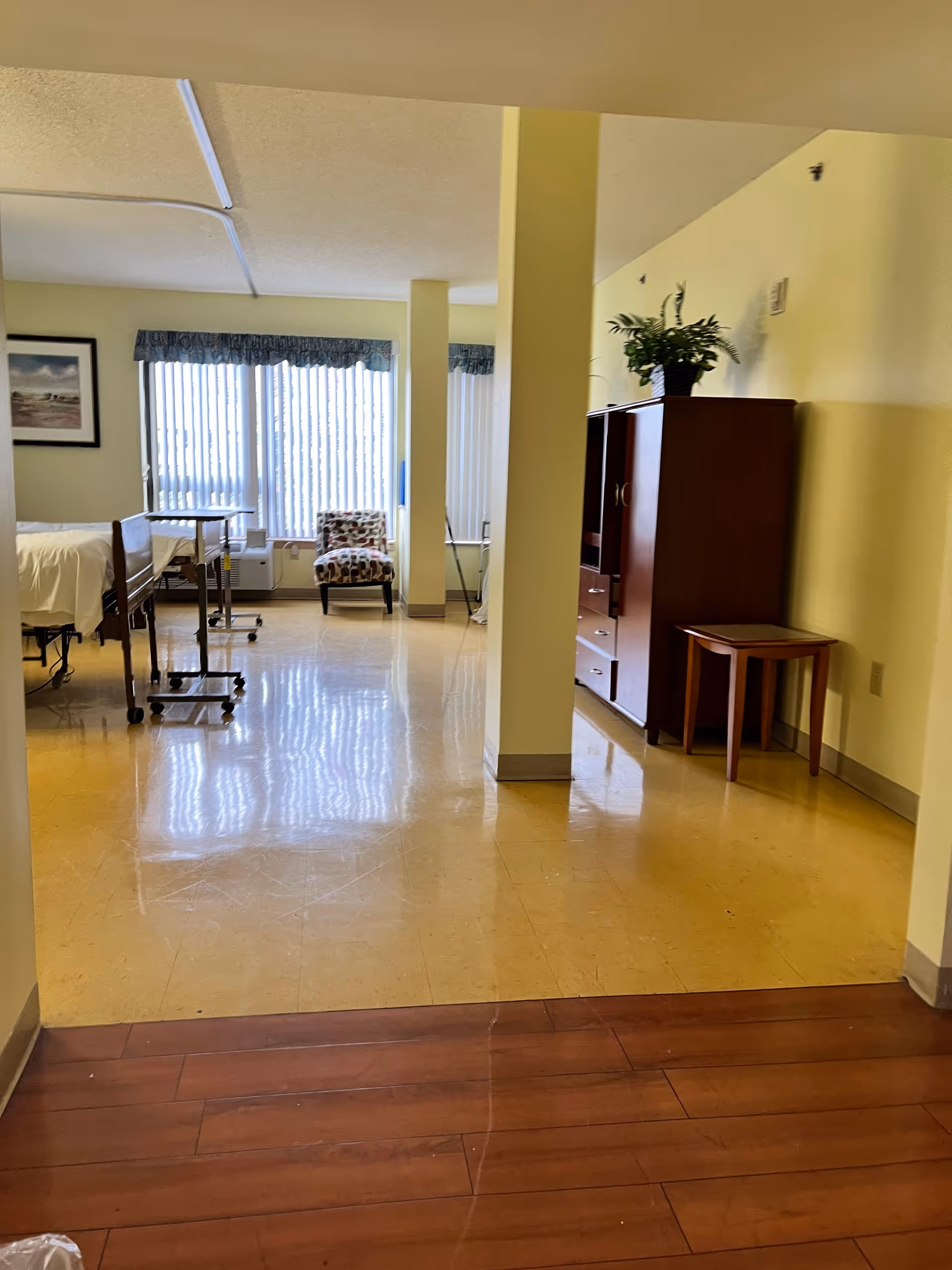 Interior of a senior living patient room with bed, chairs, cabinet, and windows.