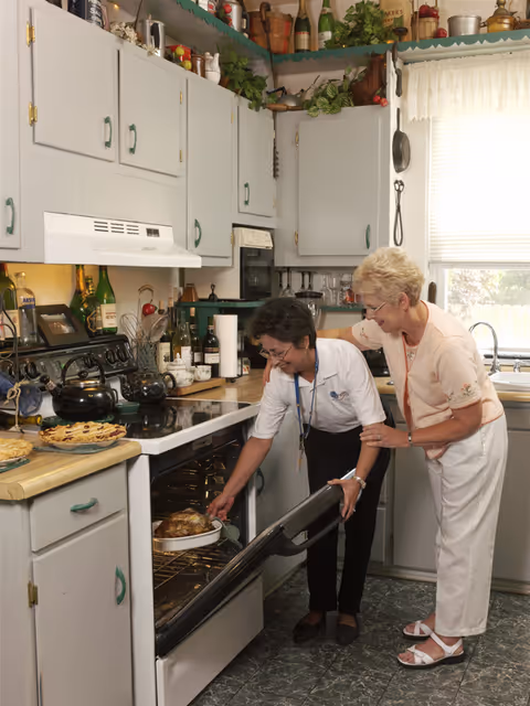 Two caregivers in a kitchen watch as a dish bakes in an open oven.