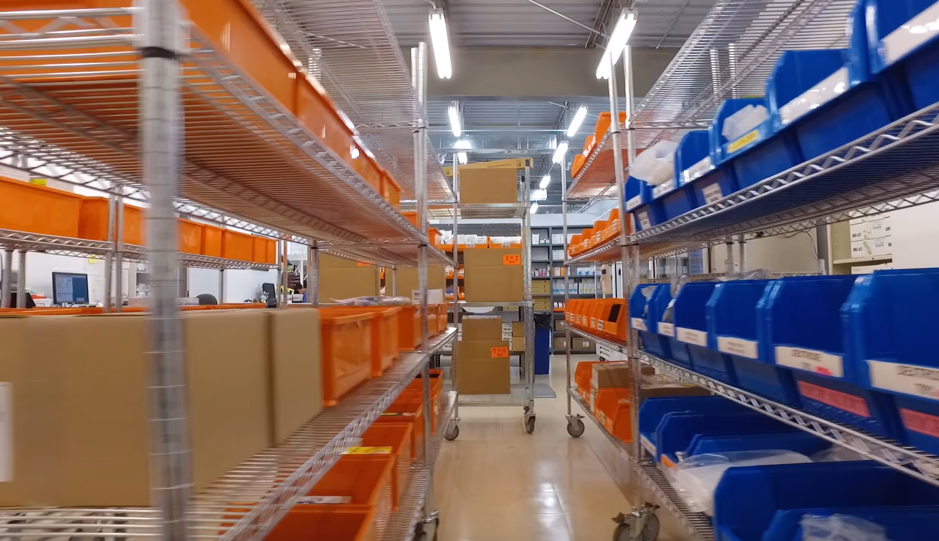 Industrial supply room with metal shelves, orange/blue bins, and stacked cardboard boxes.